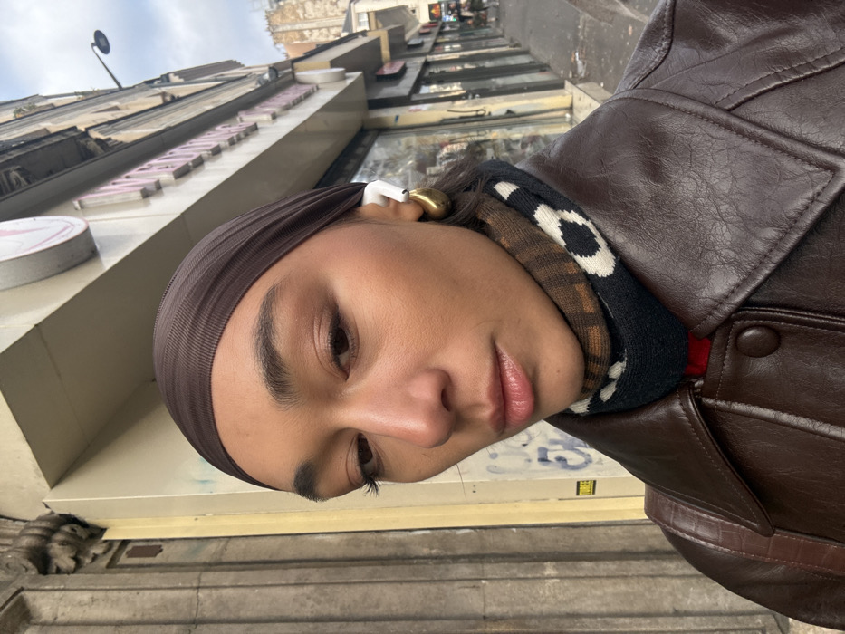 A person with a medium-dark skin tone is depicted in a close-up, slightly angled portrait. The individual wears a dark brown, ribbed head covering, a dark brown collared jacket, and a patterned turtleneck or scarf featuring brown, black, and white sections. A white wireless earbud is visible in the right ear, along with a large, gold-toned, teardrop-shaped hoop earring. A dark brown strap, possibly from a bag, rests on the left shoulder. The person maintains a neutral expression, with eyes looking slightly downwards and to the left of the frame.
The background reveals a typical Parisian street scene. Multi-story buildings constructed from light-colored stone, displaying classic architectural details including ornate carvings, line the street. Below these structures are various shopfronts with glass display windows and visible signs, one with partially obscured pink lettering. Faint graffiti is visible on a lower wall section to the left. A black, curved streetlight is positioned against a cloudy sky in the upper right. The distinct architectural style and urban elements confirm the location as a street in Paris, France.