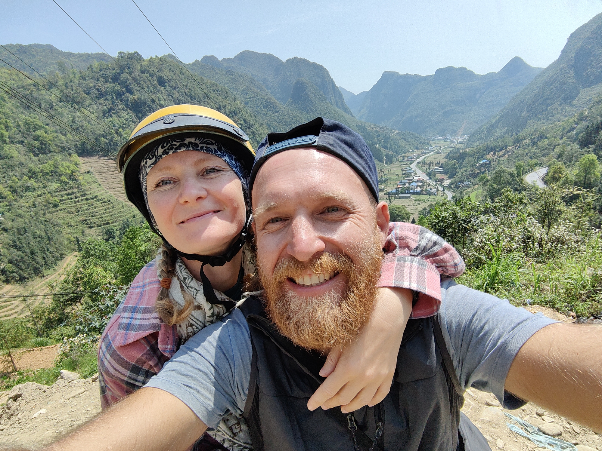 A selfie captures two individuals against a backdrop of Phố Bảng Commune, Vietnam's mountainous terrain. A woman dons a yellow helmet with a floral band beneath, her arm draped across the shoulders of a smiling man wearing a baseball cap. He has a prominent red beard. Behind them, a village nestles in the valley, bisected by a winding road. The verdant mountains stretch into the distance under a clear sky.