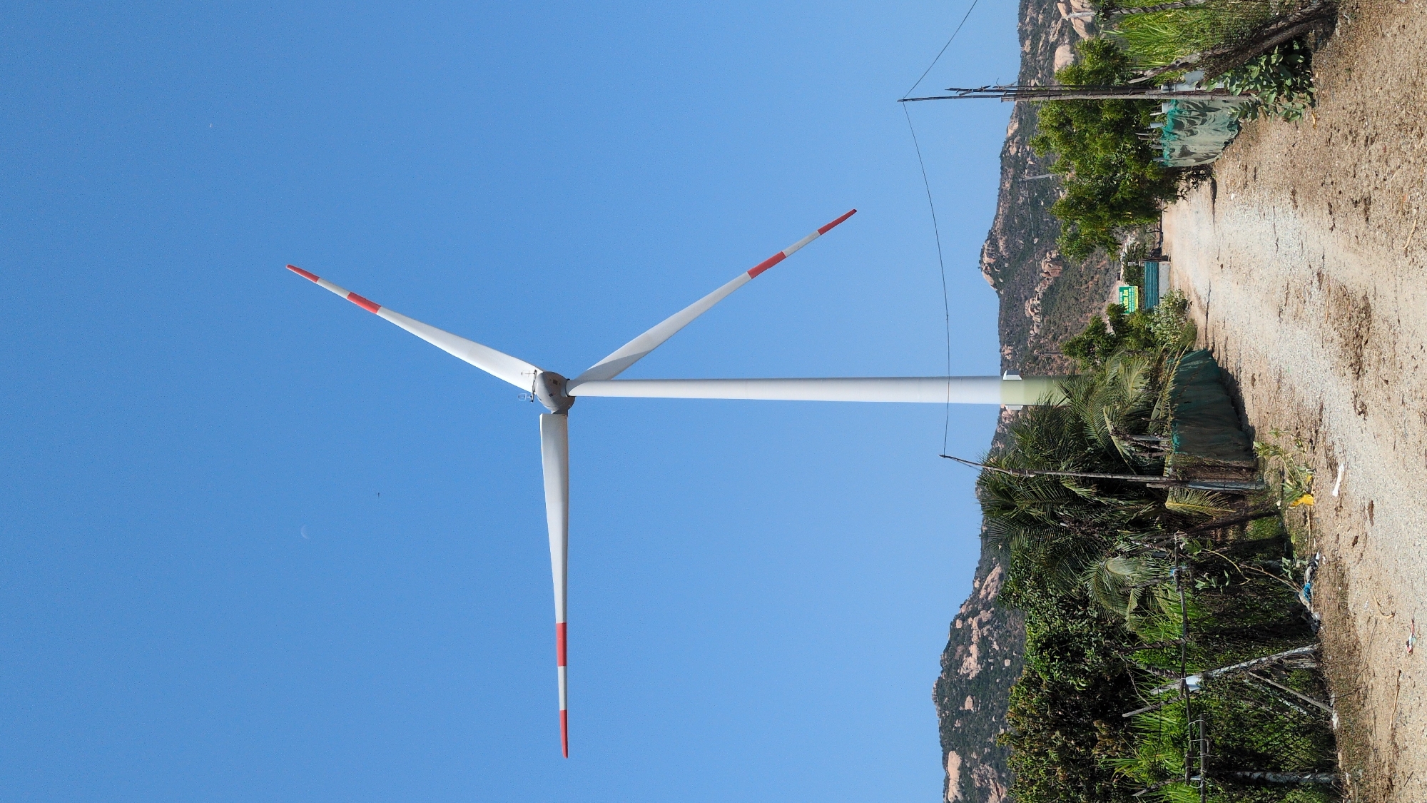 The image shows a wind turbine standing on a dirt road in Phước Dinh, Vietnam. The turbine has three white blades with red tips against a clear blue sky. Vegetation and rocky hills line the side of the road. A utility pole with wires is visible near the turbine.