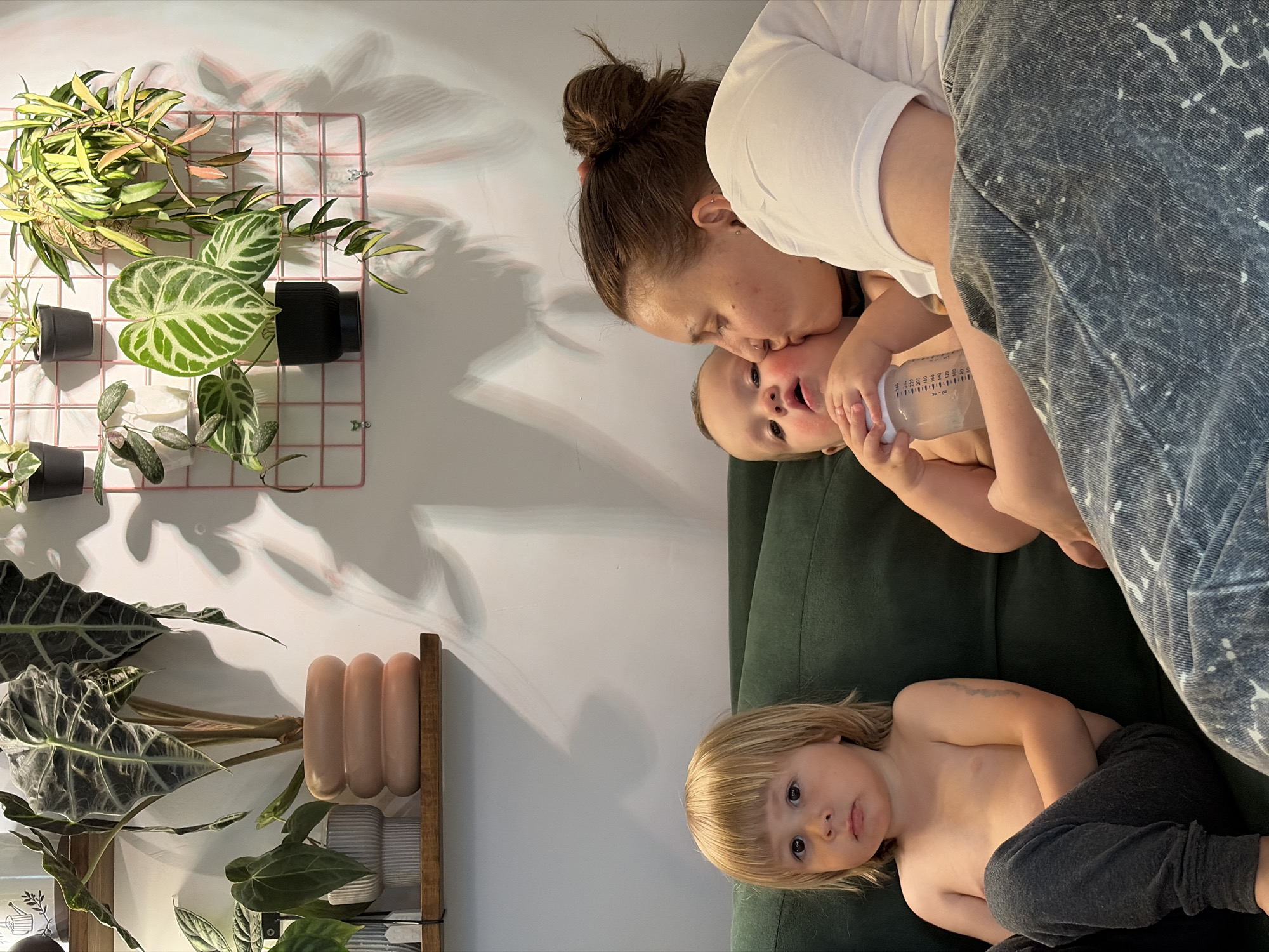 In Malyi Bukor, Russia, a family is captured indoors. A woman with her hair pulled back in a bun is kissing a baby on the cheek while holding a bottle. Beside them sits a young boy with blond hair. The family is on a dark green sofa. The background features a white wall with shelves containing various potted plants. The lighting suggests it is daytime.
