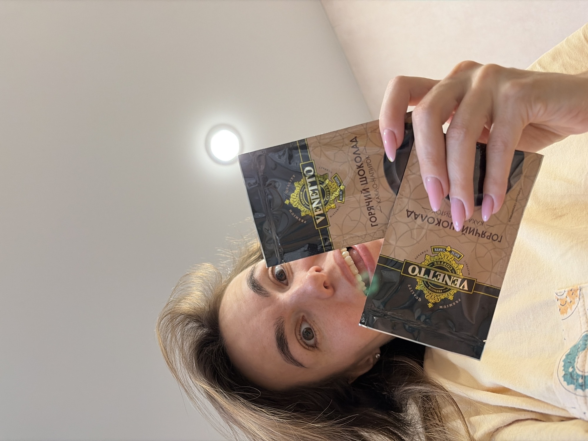 A woman is holding two packets of "Venetto" hot chocolate mix. The packaging includes Russian text. The woman is fair-skinned with brown hair and light pink fingernails. The photo is taken from a low angle, with a white ceiling and a light fixture visible in the background. Based on OCR text, the product is a hot chocolate drink mix and has the words "Izhevsk, Russia" printed on the package.