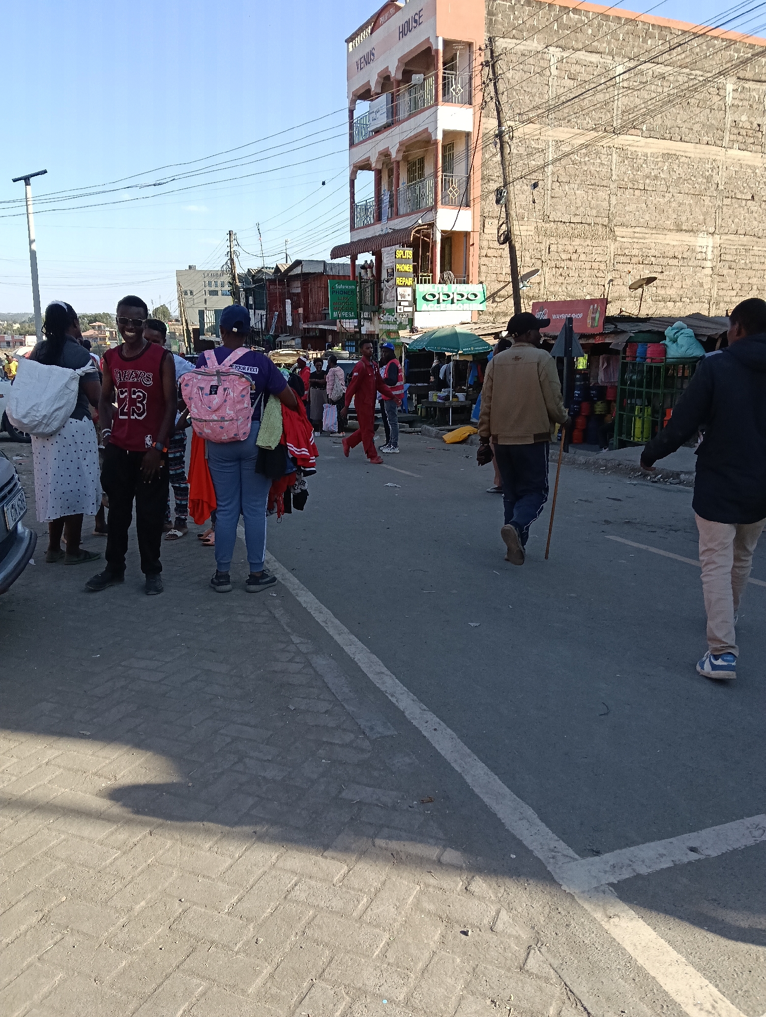 In a street scene in Keekonyokie Ward, Kenya, people are walking on a paved road. A young man in a red Lakers jersey and black pants stands prominently, smiling at the camera. Beside him, a young woman with a pink backpack and light blue jeans is seen from behind, holding several items of clothing. In the background, a multi-story building with the name "VENUS HOUSE" is visible, along with market stalls and various advertisements. The environment appears to be an urban commercial area with a clear blue sky overhead.