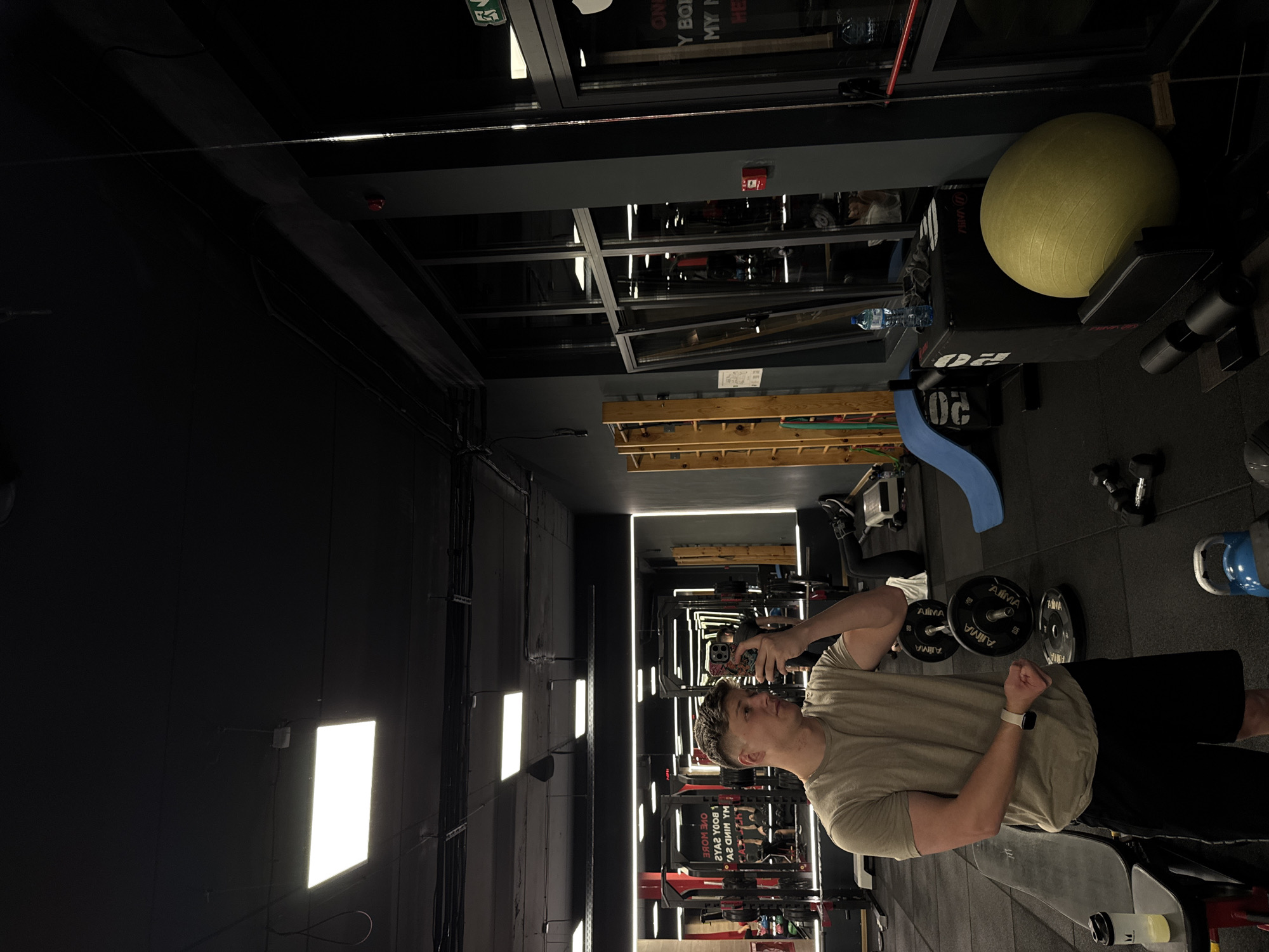 The image shows a man taking a selfie in a gym. He is wearing a beige t-shirt, black shorts, and a smartwatch. He is holding a phone with a patterned case. The gym is dimly lit, with rows of exercise equipment visible in the background. There are weights, a large exercise ball, and other fitness items in the foreground. The environment appears modern and well-equipped. It is difficult to verify the location within Plovdiv, Bulgaria, without further information.
