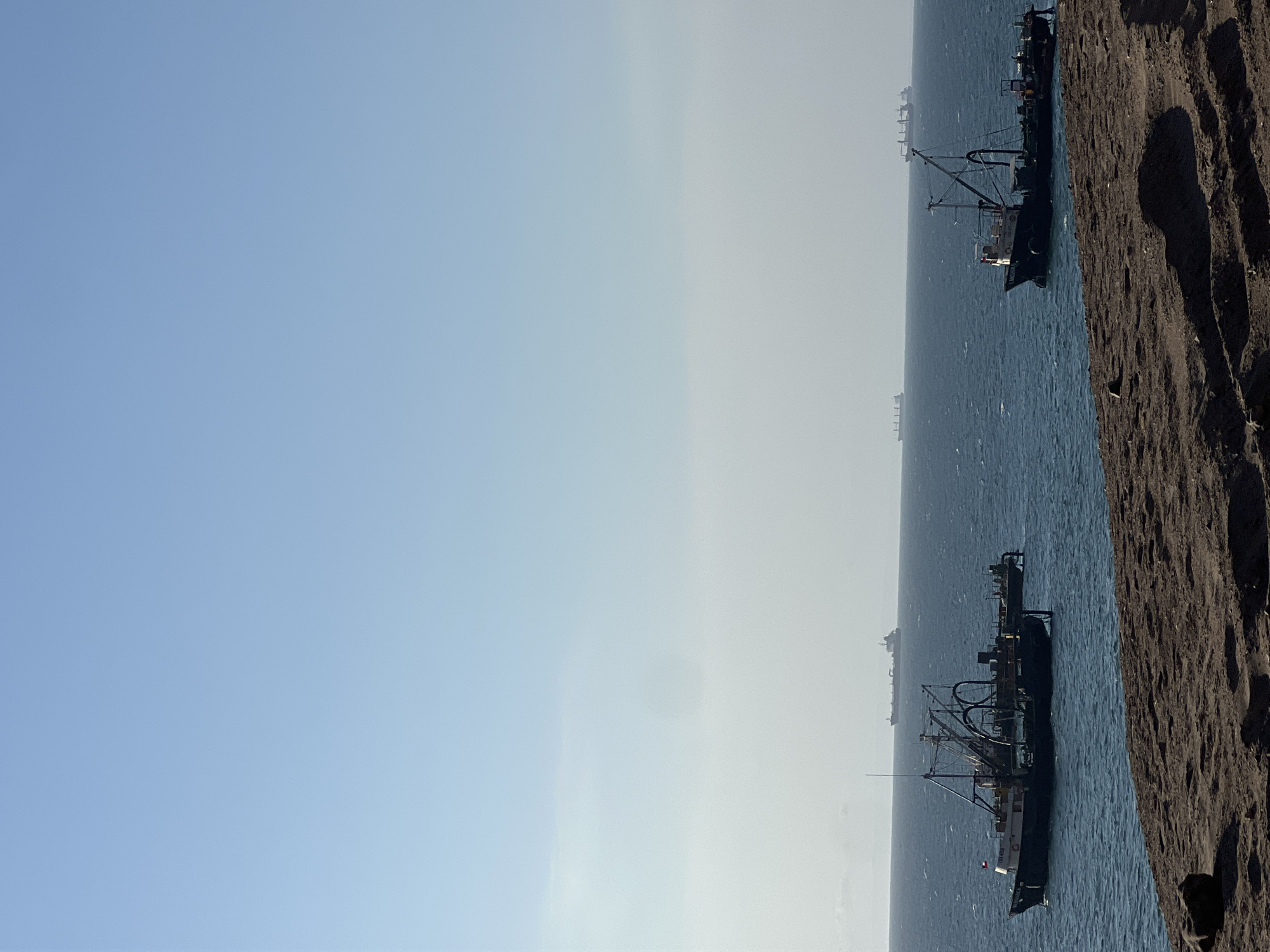 The scene depicts the coast of Arica, Chile, featuring several fishing vessels anchored near the shore. A section of dark, rocky coastline occupies the right edge of the image. To the left, the ocean stretches toward the horizon under a clear sky, with some hazy conditions obscuring the distant ships. Two fishing boats are prominent in the foreground, showing their rigging and deck equipment. In the distance, smaller vessels can be seen, blending into the horizon.