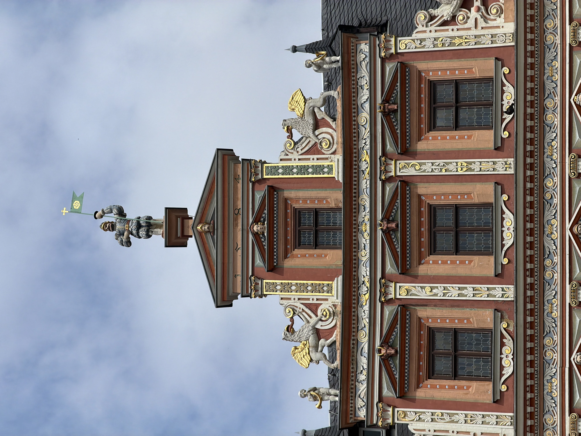 The image shows the facade of a historic building, likely the Haus zum Großen and Kleinen Horn, in Erfurt, Germany. The architecture is ornate, with a red and gold color scheme. At the very top, a statue of a man in armor holds a green flag with a golden cross and a wheel design. Below this, on the roofline, are two gilded griffin statues, each with a small cherub blowing a horn on either side. The building features multiple windows with dark panes, framed by decorative elements. The sky above is overcast with some clouds.