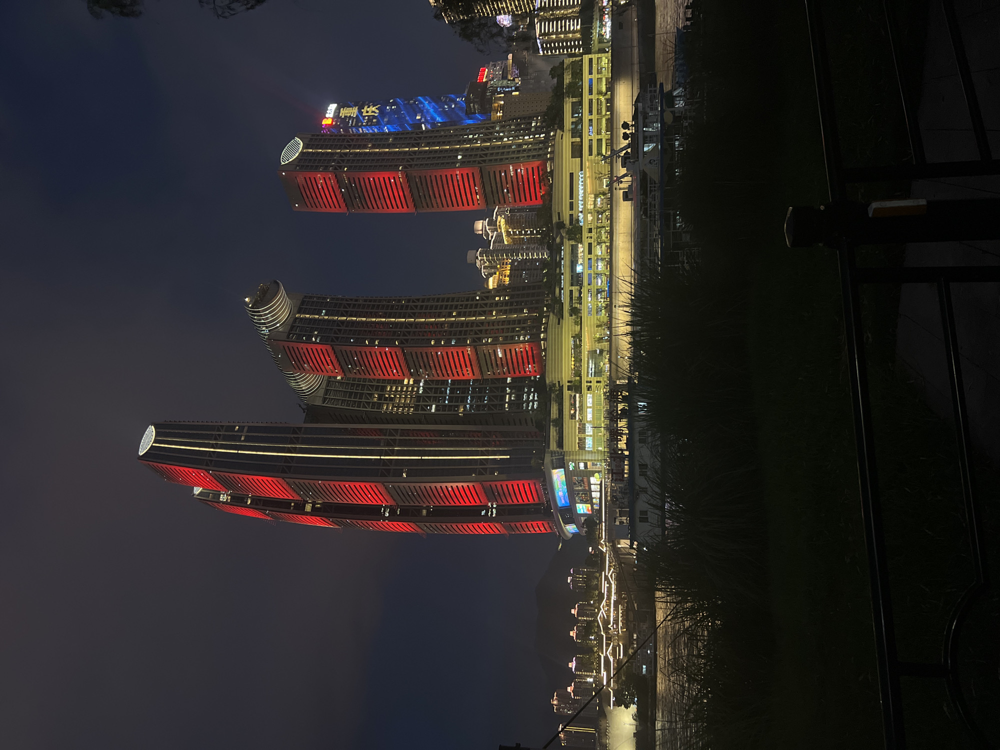 In the Yuzhong District of China, a nighttime cityscape is dominated by a cluster of illuminated skyscrapers situated along a waterfront. The most prominent structures are three tall, modern buildings with distinctive architectural features. The two tallest buildings are rectangular with rounded tops and are extensively lit with alternating bands of vibrant red and warm yellow lights, highlighting their verticality. To the right of these, another skyscraper features a predominantly red illumination. Further to the right, a slightly shorter building displays a vertical blue light pattern, and on its upper portion, a sign with Chinese characters is visible, illuminated in yellow.
Below the skyscrapers, a multi-story structure with horizontal banding of lights stretches across the mid-ground, appearing to be a commercial or entertainment complex. Several boats are docked or moving on the wide river in front of this complex. The river reflects the lights from the city and the boats, creating shimmering patterns on its surface.
In the foreground, a strip of dark grass and tall reeds obscures the lower portion of the scene, suggesting a park or promenade area along the riverbank. A dark metal railing runs horizontally across the bottom of the frame. The sky above is dark and overcast, with a faint glow emanating from the city lights. The overall impression is one of a dynamic and brightly lit urban environment at night.