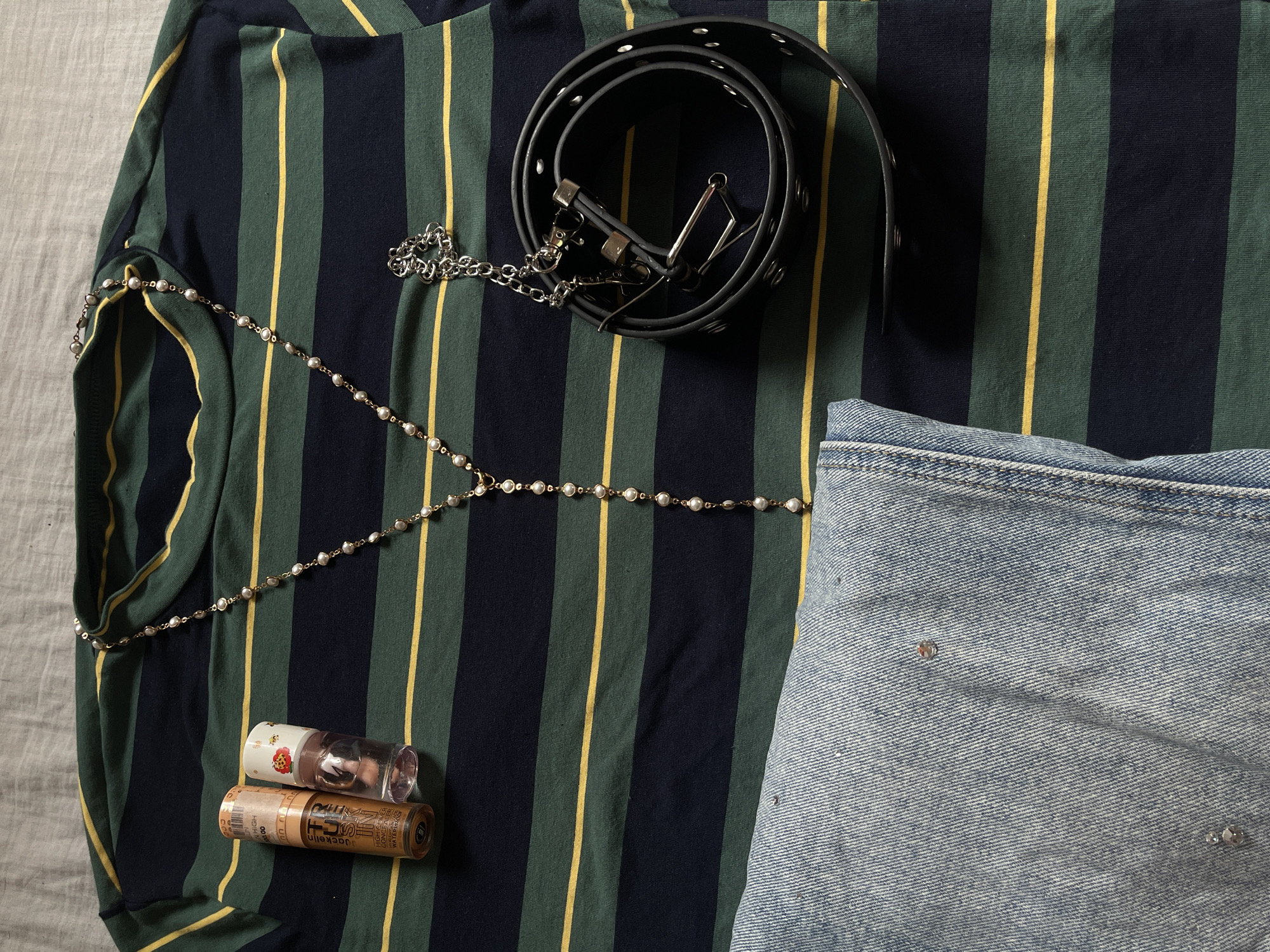 This flat lay image captures an arrangement of clothing and accessories, possibly an outfit for display, against a light grey fabric background.
The central item is a long-sleeved shirt or top featuring a bold vertical stripe pattern in navy blue, dark green, and yellow. On top of the shirt, a black studded belt with a silver buckle is coiled. A silver chain with multiple links is attached to the belt, extending towards the left. A delicate gold-colored body chain or long necklace, adorned with small white pearls, is draped across the shirt, partially visible on the left side and extending to the right.
To the right, a section of light blue denim jeans is visible, showing a classic denim weave and subtle wear. The jeans are embellished with small, scattered, reflective studs or rhinestones.
In the lower-left corner, resting on the striped shirt, are two cylindrical cosmetic tubes. The top tube is clear plastic with a small floral pattern on its cap. The bottom tube is golden-brown. Visible text on the golden-brown tube includes "Jackelin," "WATERPROOF," and "CONCEALER." A sticker label partially reveals "RM 10.00," indicating a price in Malaysian Ringgit.
The scene depicts ready-to-wear items, possibly for an outing in Ado, Nigeria, though specific local cues are absent from the objects themselves beyond the general style of attire.