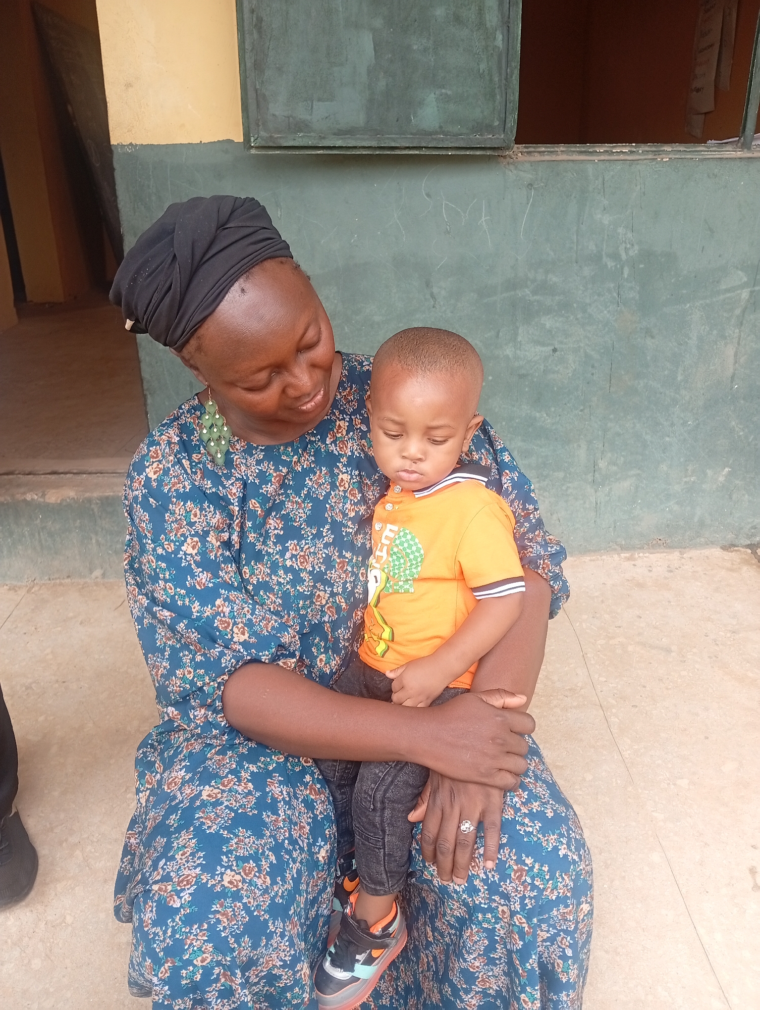 This full-length portrait captures an adult female lovingly holding a young male child on her lap. The woman, with dark skin, wears a black headwrap and a blue dress adorned with a small floral pattern in brown, orange, and green. She also sports long, light green beaded earrings and a ring on her left ring finger. Her gaze is directed downwards towards the child, with a gentle smile suggesting affection.
The child, also with dark skin and short, buzzed hair, is dressed in an orange short-sleeved polo shirt featuring a geometric pattern in green and white, along with partially visible white text on the front. He wears dark jeans and black sneakers with orange accents and light-up soles. The child is looking downwards, appearing calm.
They are seated on a light-colored paved surface, likely concrete or tiles. The background consists of a two-toned wall; the upper section is a light yellow or beige, while the lower section is a dark green. Faint white scribbles are visible on the green wall. Above and to the left, a doorway or opening reveals a darker interior space, and above the green wall, a dark green window frame or shutter is visible, with an opening above it showing another dark interior.
The bright, natural lighting indicates it is daytime. The scene evokes a tender and protective moment between the two individuals in what appears to be a casual outdoor or semi-outdoor setting in Narayi, Nigeria.