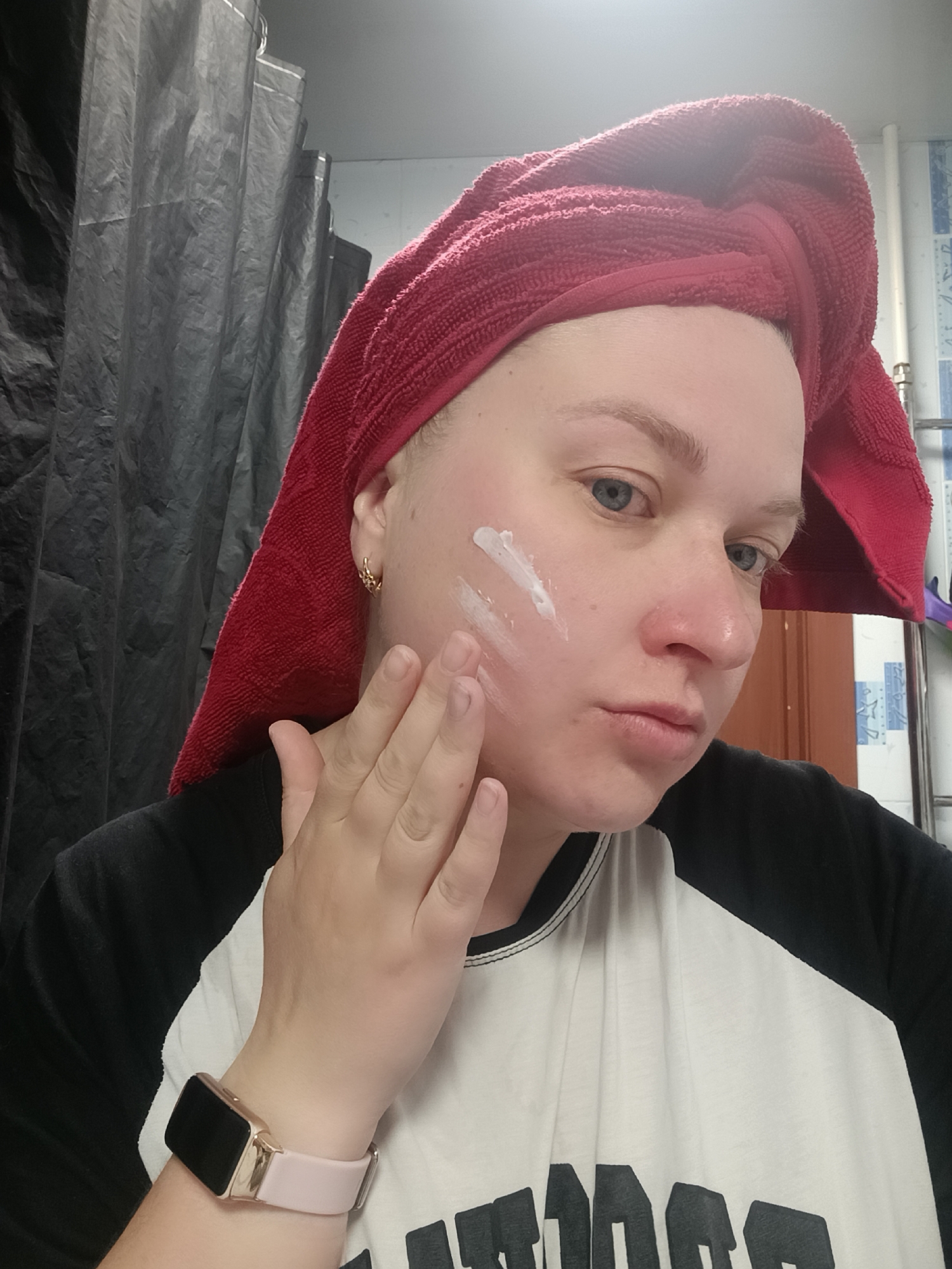 A woman is applying moisturizer to her face in a bathroom. She has a red towel wrapped around her head and is wearing a black and white shirt and a pink smart watch. White cream is visible on her cheek, which she is spreading with her right hand. The bathroom has a black shower curtain and blue and white tiled walls. The image was taken in Chaikovsky, Russia.