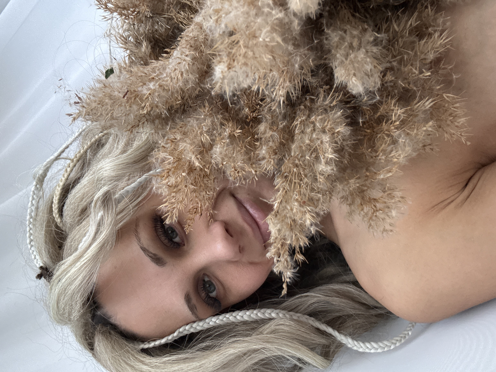 A woman with blonde hair and braided accents lies against a white surface, likely fabric or a table. A large bundle of pampas grass obscures part of her face and shoulder. The scene is well-lit. There is no verifiable information to conclude that the image was taken in Rusanovo, Russia.