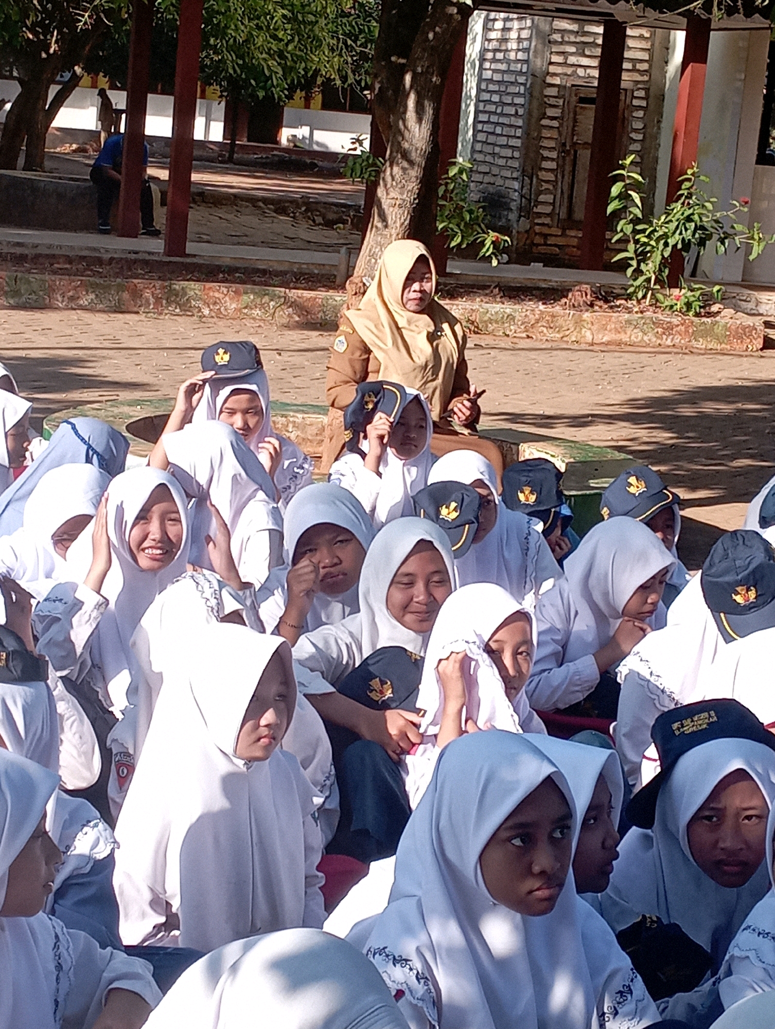 In the city of Banyuurip, Indonesia, a group of students, predominantly young girls wearing white hijabs and some wearing dark caps with yellow emblems, are gathered outdoors in what appears to be a schoolyard. The students are seated on the ground, facing in various directions. Many of them are looking towards a woman standing in the background. The woman is wearing a light brown hijab and a matching uniform, suggesting she is a teacher or school staff member. She is standing behind the seated students, holding her left hand up, possibly addressing the group or giving instructions. The environment is sunny with trees and a brick building visible in the background. The ground is paved with interlocking bricks.