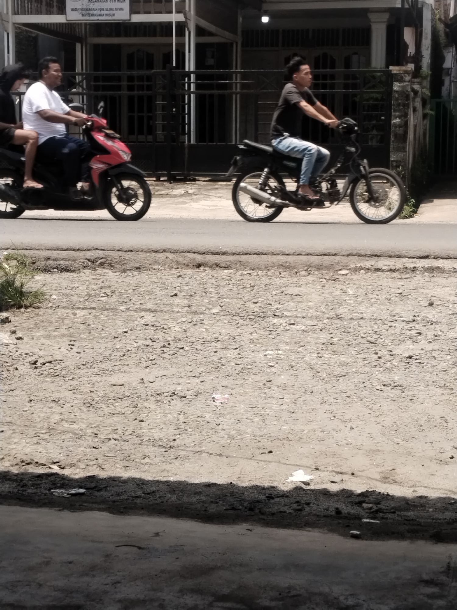 The image shows a street scene in Talun Kenas, Indonesia. Two men are riding motorcycles. In the foreground, a man in a white shirt and dark shorts rides a red and black scooter. He has a passenger behind him wearing dark clothing. In the background, a man in a black t-shirt and blue jeans rides a black motorcycle. The motorcycles are on a paved road. The road is bordered by a gravel shoulder on the right. A building with a dark fence is visible behind the motorcycles. The sky is bright, indicating daytime.