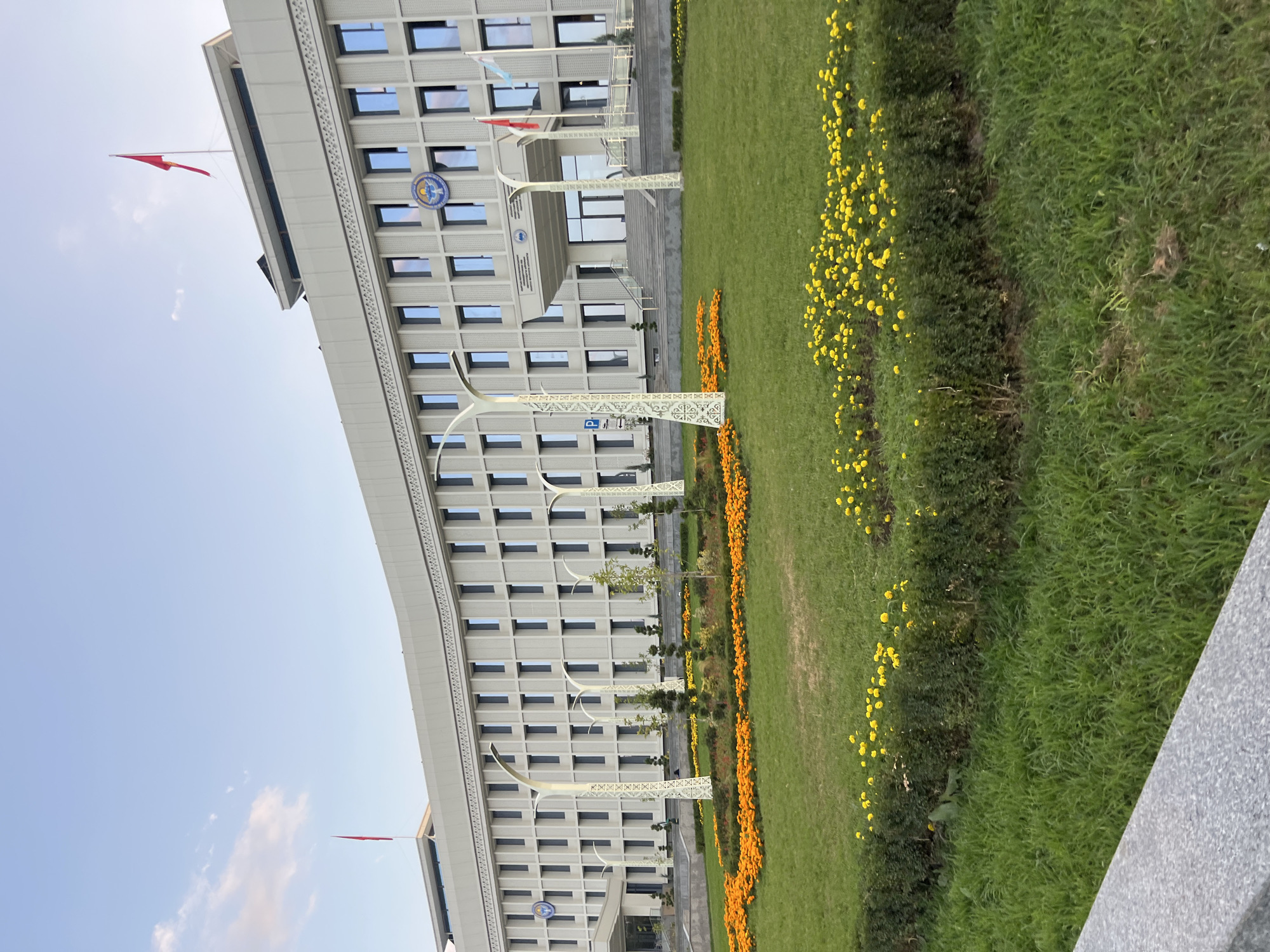 The image shows a modern, light-colored, multi-story building with a curved facade. The building has numerous rectangular windows and decorative architectural elements. Several flags are visible on poles on the roof and near the entrance. In front of the building is a well-maintained lawn with flower beds filled with bright orange and yellow flowers. There are also several ornate lampposts and some trees. The sky is blue with a few wispy clouds. The image appears to have been taken during the day. Based on the building's architecture and the presence of national flags, this is likely a government or official building. Without additional context or identifying markers within the image, pinpointing the exact location within Osh City is not possible.