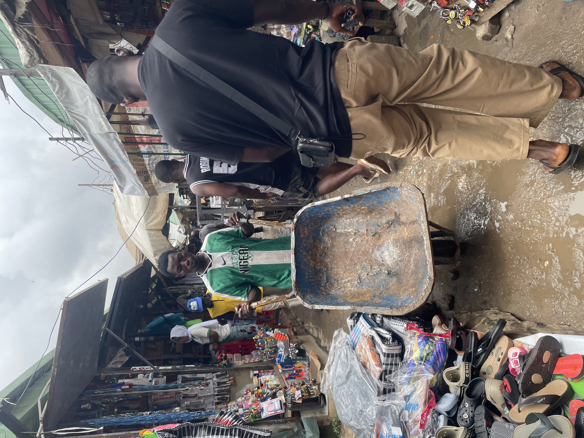 In a bustling open-air market in Abuja, Nigeria, a young man wearing a green and white Nigeria soccer jersey and a blue cap pushes a rusty blue wheelbarrow through a muddy, wet pathway. The market is lined with stalls displaying various goods, including tools, locks, and clothing. In the foreground, a pile of colorful footwear, including flip-flops and sandals, is visible. Behind the wheelbarrow pusher, another man in a black t-shirt and khaki pants walks away from the camera, carrying a black crossbody bag. The overall environment suggests a busy commercial area with a recent rainfall, indicated by the wet ground and puddles.