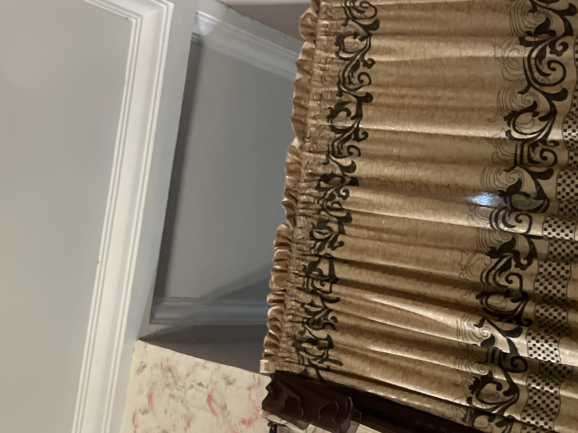 The image shows a close-up view of decorative curtains hanging in a room. The curtains are a light golden color with a textured pattern and feature a dark brown ornate scrollwork design along the bottom edge. Above the curtains, white decorative molding is visible on the ceiling, and a portion of a wall with a pink and grey patterned wallpaper is to the left. The background appears to be a plain grey wall. There are no people or discernible actions in the image. The scene is indoors, likely a residential setting. The location is not specified in Jalingo, Nigeria.