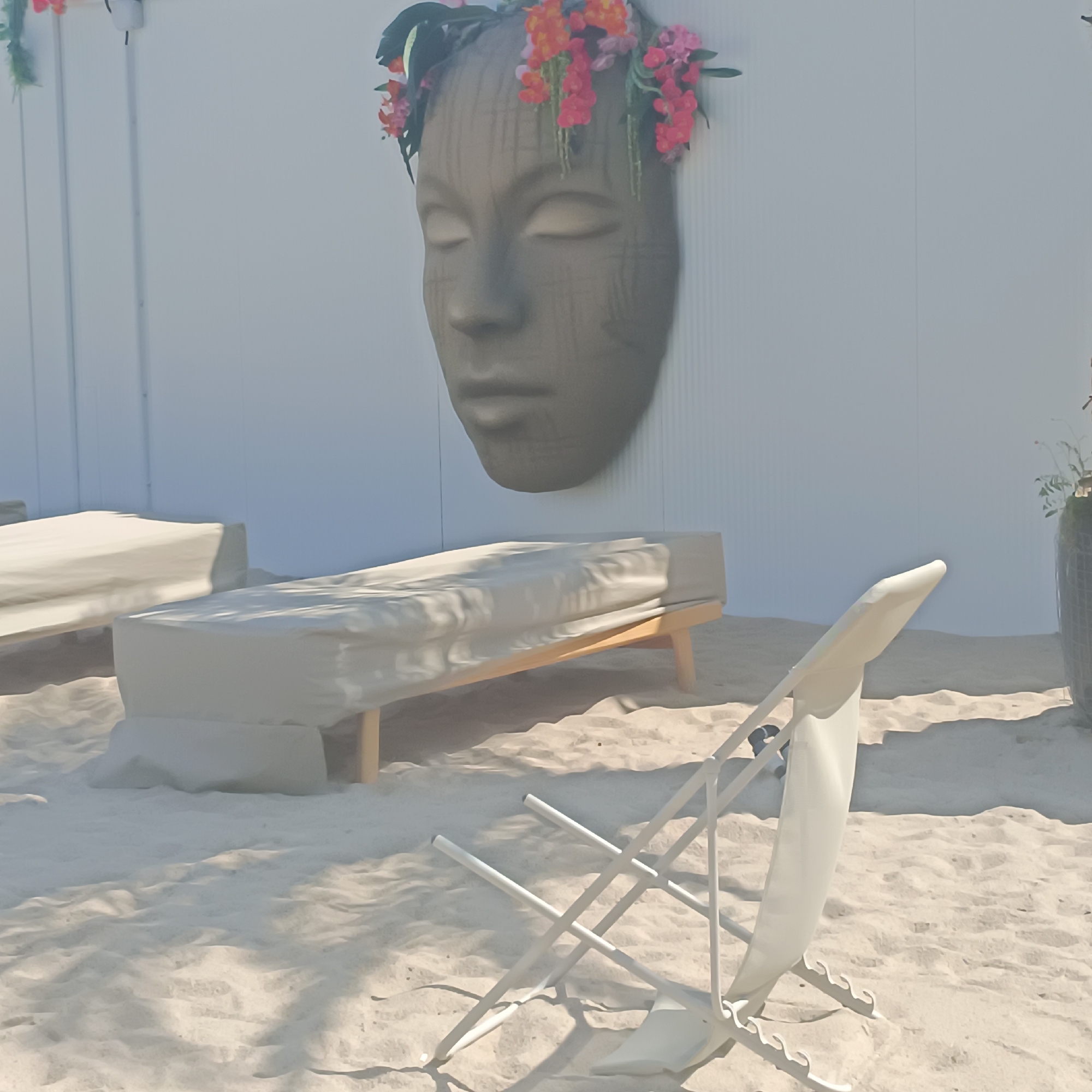 The image depicts an outdoor scene on a sandy surface, likely a beach or a courtyard. In the background, a white, corrugated wall serves as a backdrop. Mounted on this wall is a large, grey, mask-like sculpture of a face adorned with a floral wreath of pink and orange flowers. In the mid-ground, two long, flat, cushioned loungers covered in a light-colored fabric are positioned on the sand. A white, foldable beach chair is in the foreground, partially reclined and angled towards the right side of the frame. The overall impression is one of a tranquil, possibly recreational or decorative outdoor space. The image does not contain any elements that would place it specifically in Frankfurt, Germany.