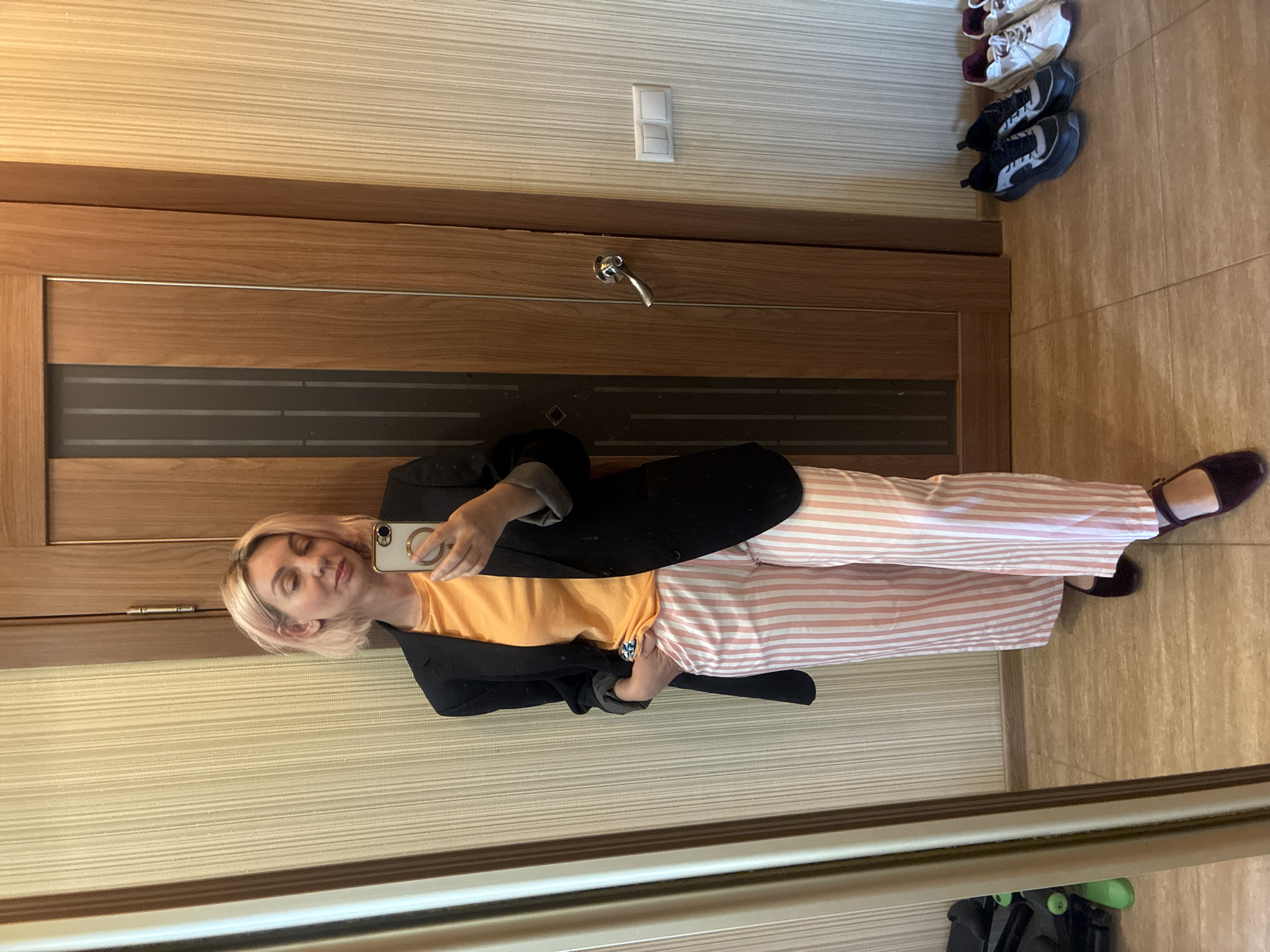 A blonde woman is captured in a mirror selfie, standing in what appears to be a residential hallway or entryway. She is wearing a black blazer over an orange t-shirt, paired with light pink and white striped wide-leg trousers, and dark flat shoes. Her eyes are partially closed, giving her a calm and relaxed expression as she holds up a smartphone, visible with a ring accessory on its case, to take the picture.
Behind her, a wooden door with dark horizontal panels and a metallic strip is visible, featuring a chrome handle. Above the door handle, on the light-colored, horizontally striped wallpaper, there is a white light switch or outlet. To the right of the door, on the tiled floor, several pairs of shoes are neatly lined up, including white sneakers with burgundy accents and black sneakers with white accents. The floor is covered with light brown tiles. The overall setting suggests a modern indoor environment. No specific time of day or weather can be determined from the image, nor are there any visible texts. The provided context indicates the location is Sochi, Russia.