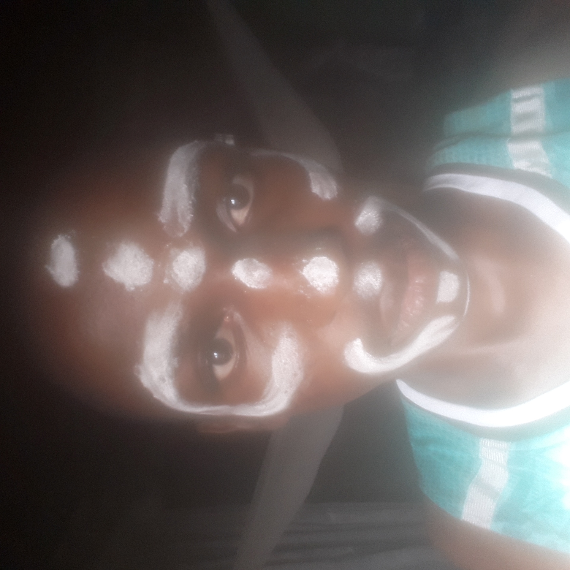 A close-up, rotated image shows a dark-skinned person with white paint or powder markings on their face. The markings consist of circular shapes on the forehead and cheek areas, and linear markings around the eyes and mouth. The person is wearing a garment with a white and teal striped pattern. The background is dark and indistinct. There is no definitive information in the image to indicate that this photo was taken in Nkpor, Nigeria.
