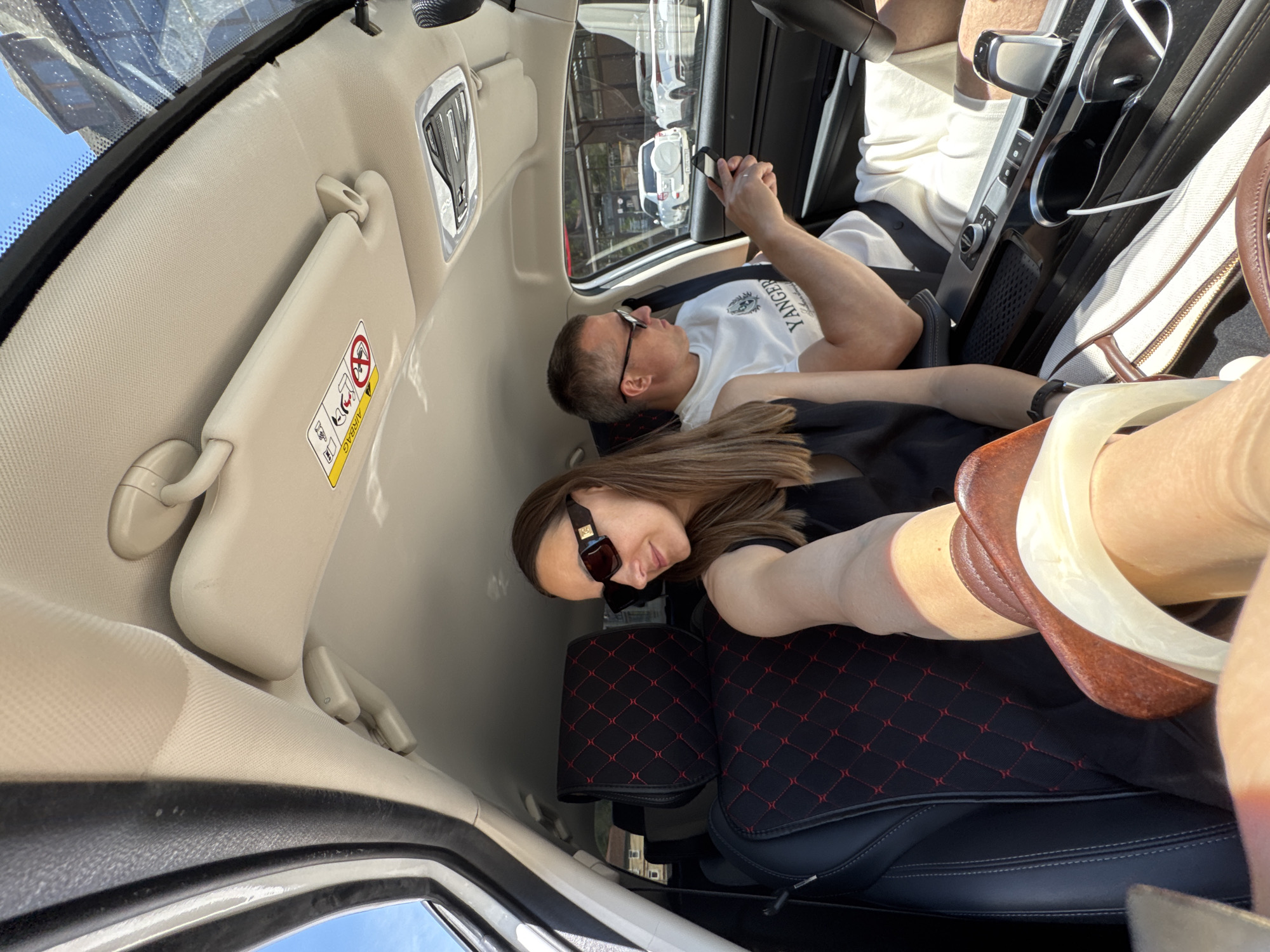 The scene is set inside the front cabin of a modern vehicle on a bright, sunny day, in what is stated to be the city of Sochi, Russia. A female individual, positioned in the front passenger seat, is capturing the image. She has long brown hair and wears dark sunglasses with gold-colored branding on the temples. Her left arm and lower leg, adorned with a brown and white open-toed sandal, are visible in the foreground. She is dressed in a dark, likely black, sleeveless top.
A male individual occupies the driver's seat. He has short dark hair and wears dark-framed sunglasses, a white short-sleeved t-shirt featuring "YANGER" and an emblem, and light-colored shorts. His right hand holds a dark smartphone, which he is observing. His left hand is partially visible on the dark steering wheel.
The car's interior features a beige fabric headliner with an overhead console housing a light fixture and controls. The front passenger sun visor is beige and displays a prominent yellow "AIRBAG" warning decal. The car seats, including the one visible on the passenger side, are black with distinctive red diamond-pattern stitching on both the headrest and seat base. The center console is dark with silver trim, incorporating a gear selector, cup holders (one containing a white cable), and other control buttons. A light-colored bag with brown leather accents is visible on the floor area between the seats.
Through the driver's side window, a clear blue sky and an outdoor urban environment are visible. Several vehicles are present: a white SUV with an externally mounted spare tire on the rear hatch, positioned in the mid-ground, and to its right, a white convertible sports car. Reflective glass buildings are also discernible in the background, suggesting a modern commercial or residential area.