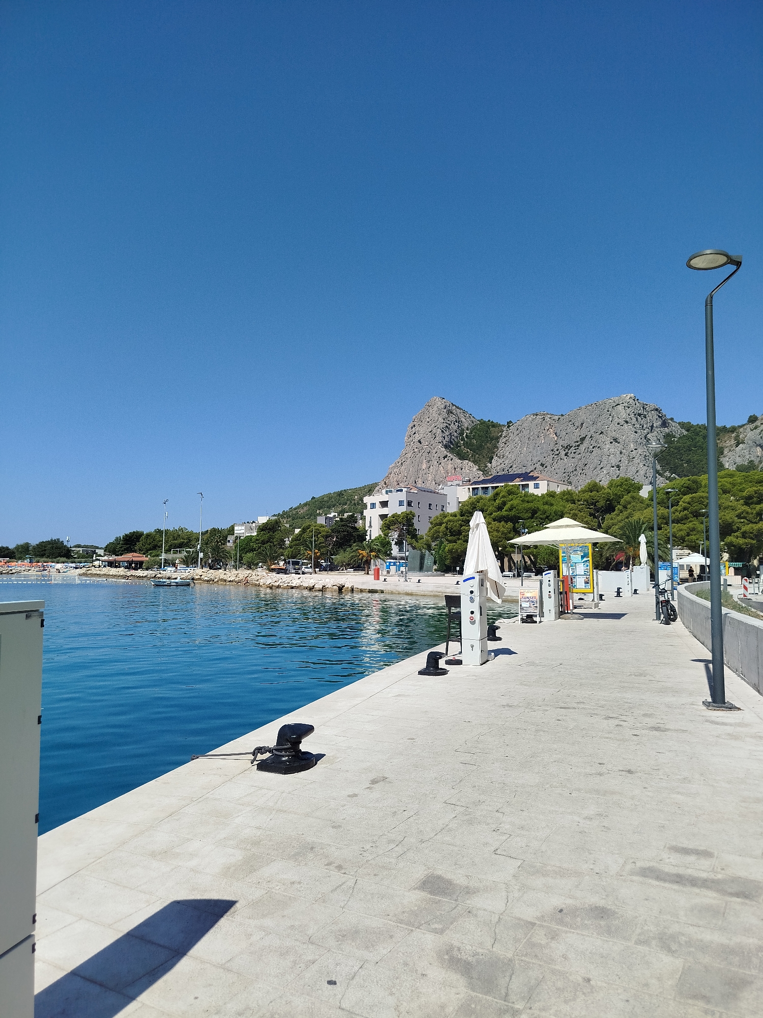The image shows a sunny day on a concrete promenade in Omiš, Croatia, overlooking the Adriatic Sea. The promenade is lined with bollards and streetlights. In the background, there are buildings, trees, and a rocky mountain. Several boats are docked in the harbor, and people can be seen walking along the promenade and on the beach. The water is a vibrant blue, and the sky is clear and cloudless. The scene conveys a peaceful and idyllic atmosphere.