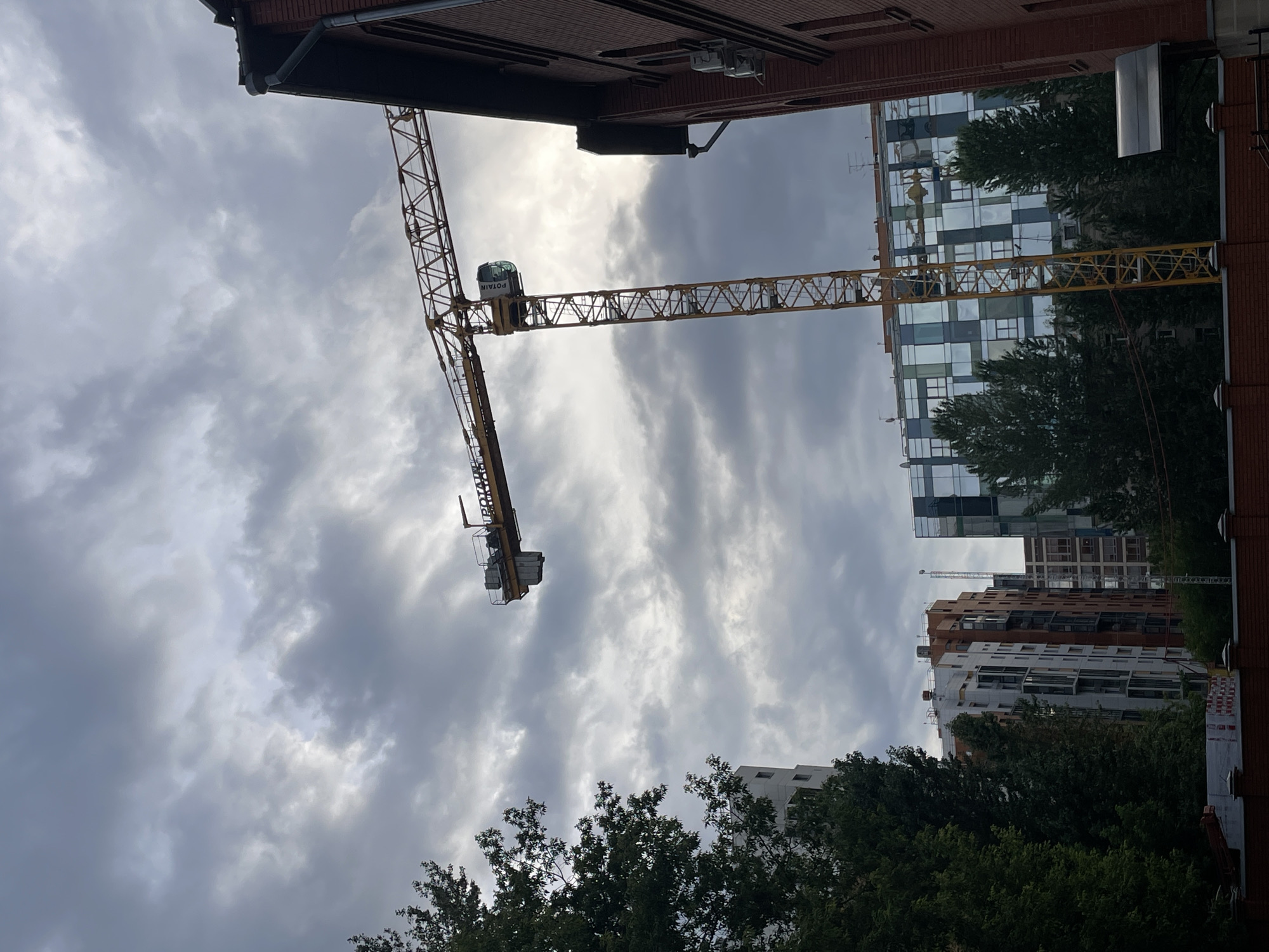 A tall, yellow construction crane is the central focus of the image, positioned against a dramatic, overcast sky. The crane's boom extends horizontally across the upper portion of the frame, with its cab visible near the main vertical structure. To the right of the crane, a portion of a red brick building with a dark, sloped roof and a visible drainpipe is prominent. In the background, several multi-story buildings of various architectural styles are visible. One building features a facade with a grid of glass panels reflecting the sky, interspersed with colored sections. To the left, a dense cluster of dark green trees partially obscures a tall, white building. In the foreground, a red brick wall with decorative white posts runs across the bottom of the frame, partially obscured by more foliage. The scene suggests an active construction site within an urban environment.
