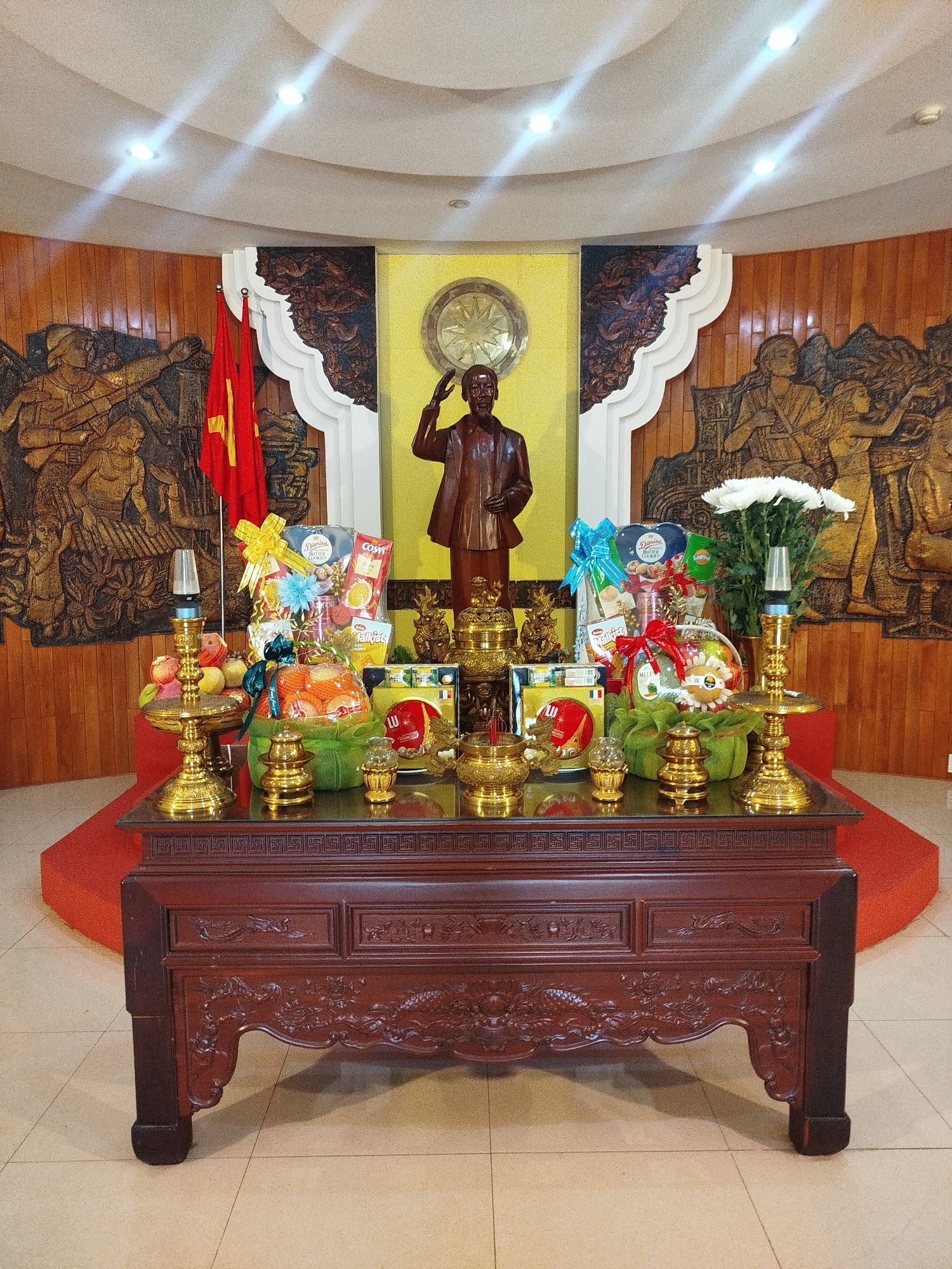 The image depicts an interior altar arrangement, likely located in Pleiku Ward, Vietnam, based on cultural objects present. A dark wood table stands on a tan tiled floor, with a red carpet visible behind it. The table is decorated with gold-colored ornaments, including what appears to be candle holders and smaller decorative pieces. There are gift baskets containing fruit and packaged goods placed on the table. Behind the table is a backdrop featuring a statue of a man in a suit saluting, presumably a historical figure. A red flag with a yellow star, the flag of Vietnam, stands to the left of the statue. Carved wooden panels flank the statue, depicting figures involved in activities such as playing music and working. The walls are paneled and a false ceiling with recessed lighting is visible.