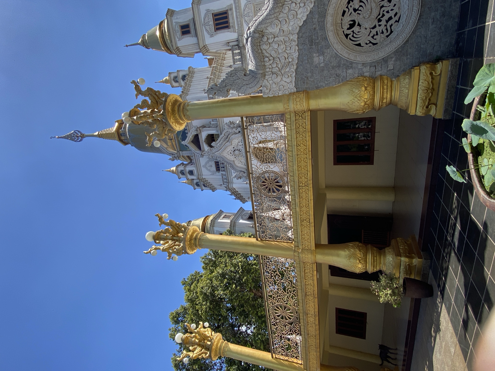 A low-angle shot showcases a Buddhist temple in Dĩ An, Vietnam. The temple exhibits a white and gold color palette. Golden pillars with decorative elements flank the temple's entrance. The temple is surrounded by a clear blue sky, a tree, and a tile pathway.