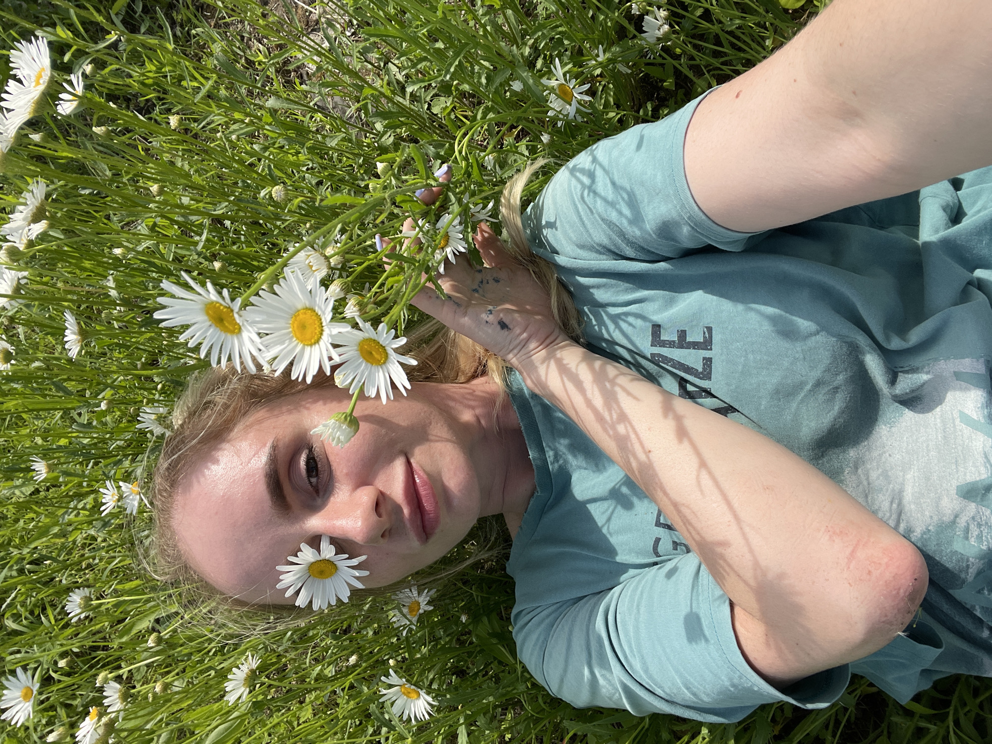 A young woman with blonde hair and daisies over her eyes lies in a field of daisies and tall green grass. She is wearing a teal t-shirt with a faded graphic on the front. Her right arm is bent, resting on her chest, and her left arm is outstretched to the side. The scene is lit by bright sunlight, casting shadows from the grass and flowers onto the woman and her clothing. The background is a blur of green grass and white daisies, suggesting an outdoor, natural setting, likely in the city of Yasnoe, Russia, given the context.