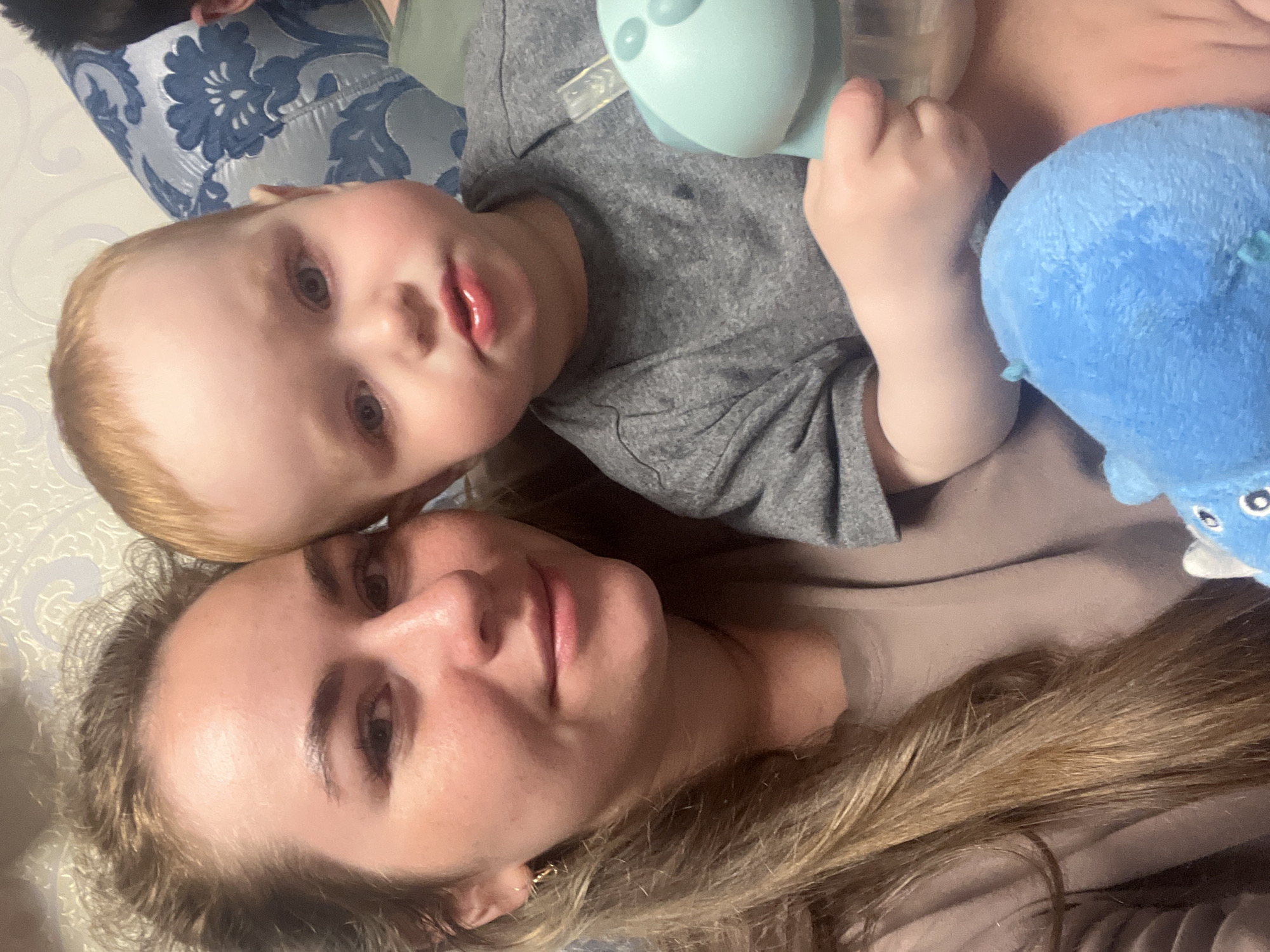 A woman and a young child are shown in a close-up selfie. The woman has long, light brown hair and is wearing a light brown shirt. The child, who has blond hair, is wearing a gray shirt and holding a light blue toy. They appear to be lying down. A blue stuffed animal is next to the child. A blue and white patterned pillow is visible in the background. The location is not identifiable based on the image.
