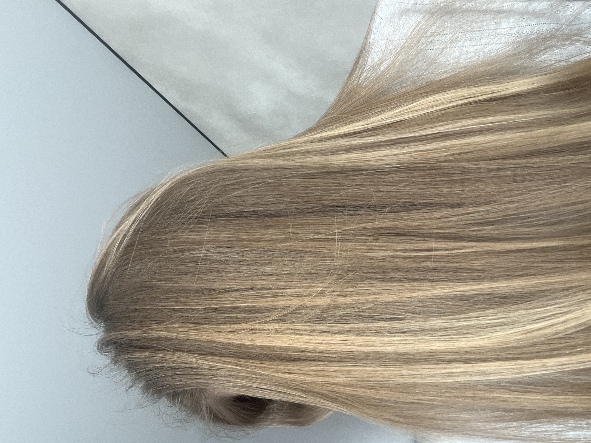 The image primarily features a close-up view of long, straight hair, likely belonging to a person, seen from a high angle or from behind. The hair displays a natural light brown or dark blonde base with distinct lighter blonde highlights or natural variations running through it. The strands appear fine and relatively smooth.
The hair is draped or lying against a light-colored background, which seems to consist of at least two distinct surfaces. In the top left portion of the image, a light blue-grey, smooth surface is visible, separated by a thin, dark diagonal line from a textured, off-white or light grey surface. This textured surface, particularly visible behind the longer strands of hair, resembles fabric, possibly a bedsheet or pillowcase.
There is no discernible activity taking place; the image focuses on the static appearance of the hair and its textures against the minimalist background. The lighting is bright and even, suggesting either daylight or well-lit indoor conditions. No specific location cues are visible within the image itself to confirm the provided context of Votkinsk, Russia, nor are there any visible emotions or text. The overall impression is one of natural hair color and texture in a clean, understated indoor setting.
