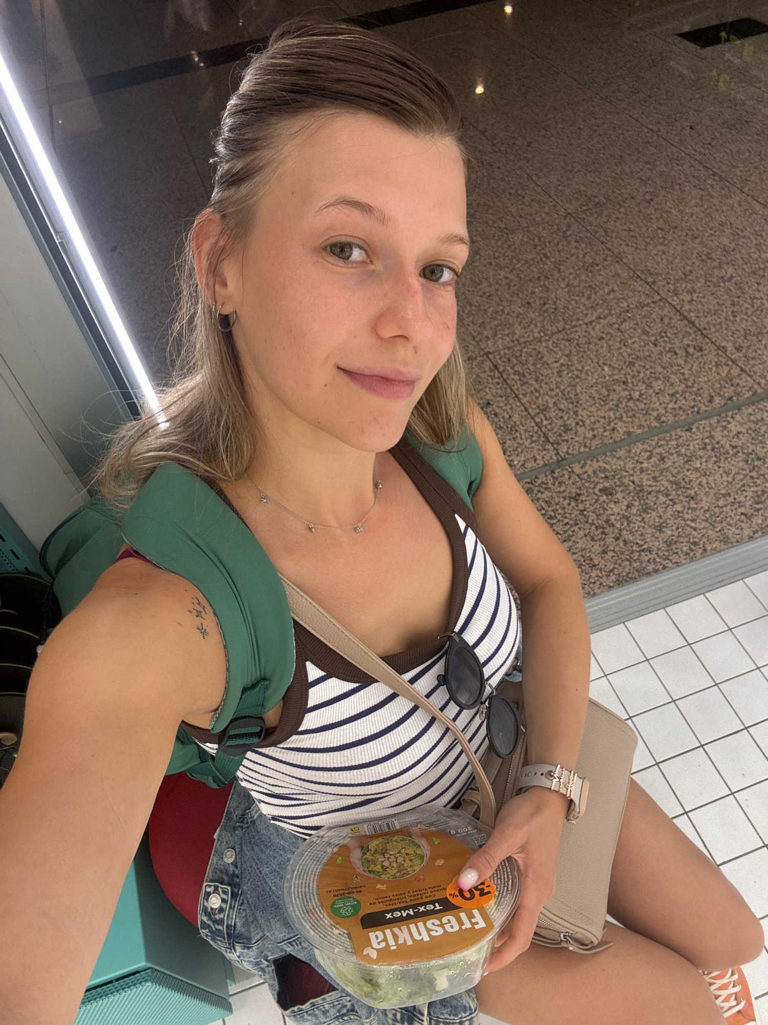 The image captures a woman posing for a selfie, holding a container of "Freshakia Tex-Mex" salad. She is seated on what appears to be a bench or low wall, with a speckled stone-like floor behind her and a white tiled floor in front. A green backpack is slung over her shoulder, and she wears a striped tank top, denim shorts, and orange sneakers. A light beige clutch purse rests on her lap. The background suggests an indoor public space, possibly an airport terminal or a train station, given the lighting and flooring. The presence of the salad container indicates she might be preparing for travel or a meal on the go. The overall impression is that of a casual moment captured in a transit hub.
