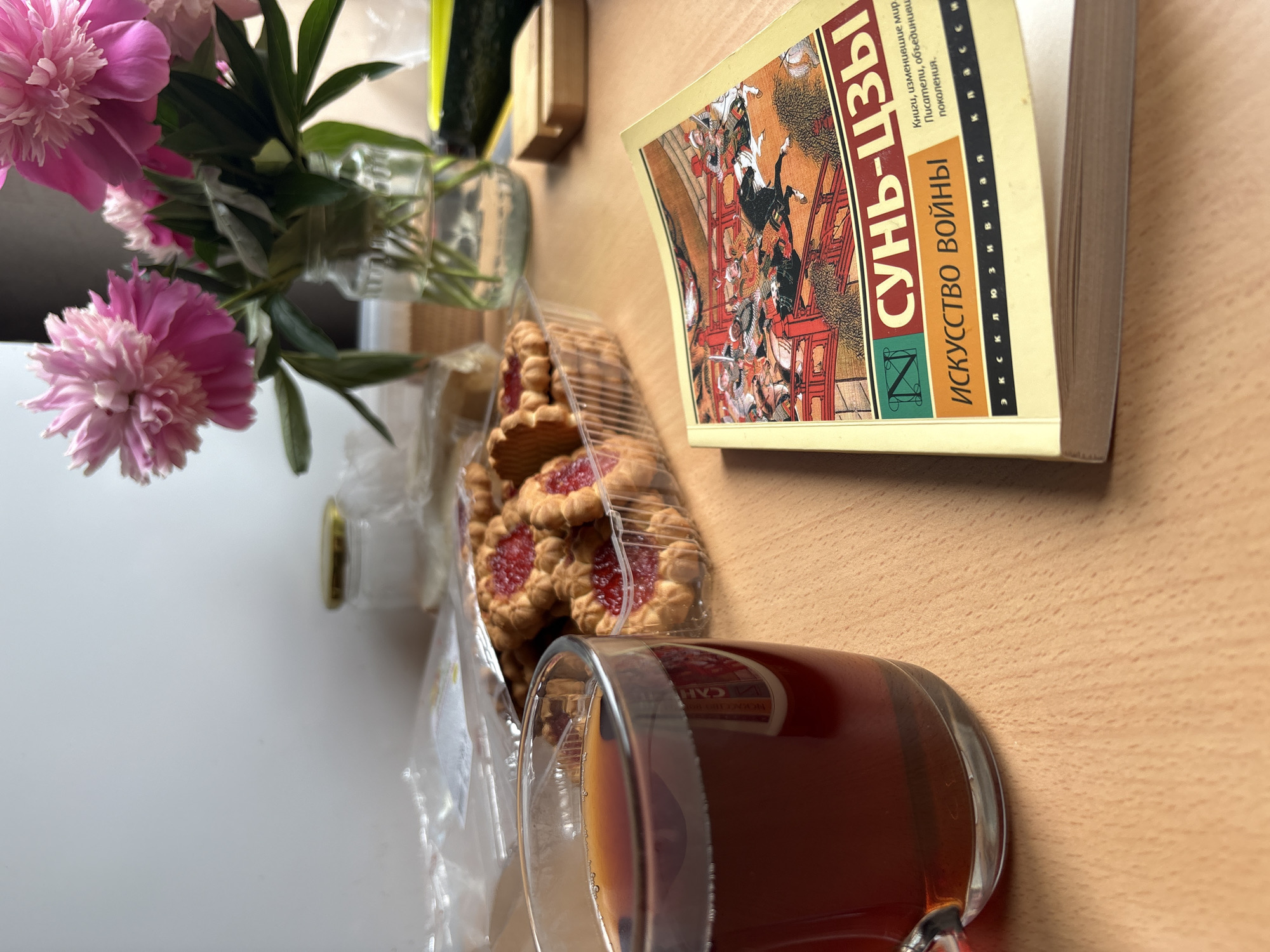 A still life photograph depicts a wooden table with several objects. In the foreground, a clear glass mug filled with dark liquid, likely tea, sits to the left. Next to it, a plastic container holds several cookies with red jam filling. To the right, a book with a yellow cover is open, displaying an illustration of people on horseback crossing a bridge. The book's title is in Russian, reading "Сунь-Цзы Искусство войны" (Sun Tzu The Art of War). In the background, a glass jar with a gold lid is visible, along with a bouquet of pink peonies in a glass vase. The overall lighting suggests an indoor setting, possibly a home or office. The location in the city of Chaikovsky, Russia is inferred from the use of the Russian language on the book cover and the general aesthetic.