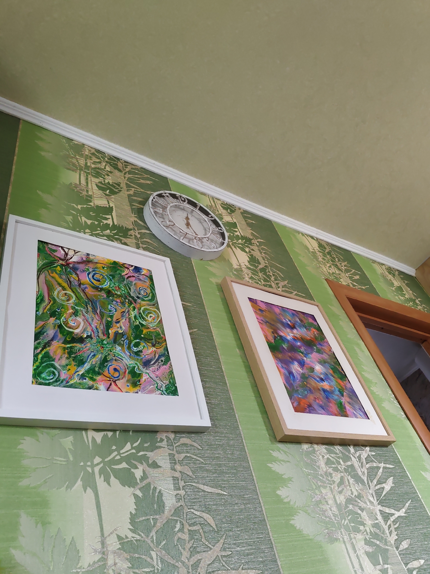 The image shows a close-up, low-angle view of a section of a wall in Rostov-on-Don, Russia, adorned with green striped wallpaper featuring a light-colored foliage pattern. Two framed abstract paintings hang on the wall, one with a white frame and the other with a light wood frame. Above the paintings, a round, white wall clock with Roman numerals is mounted. To the right of the paintings and clock, part of a wooden door frame is visible. The ceiling above the wallpaper is a plain, light beige color.