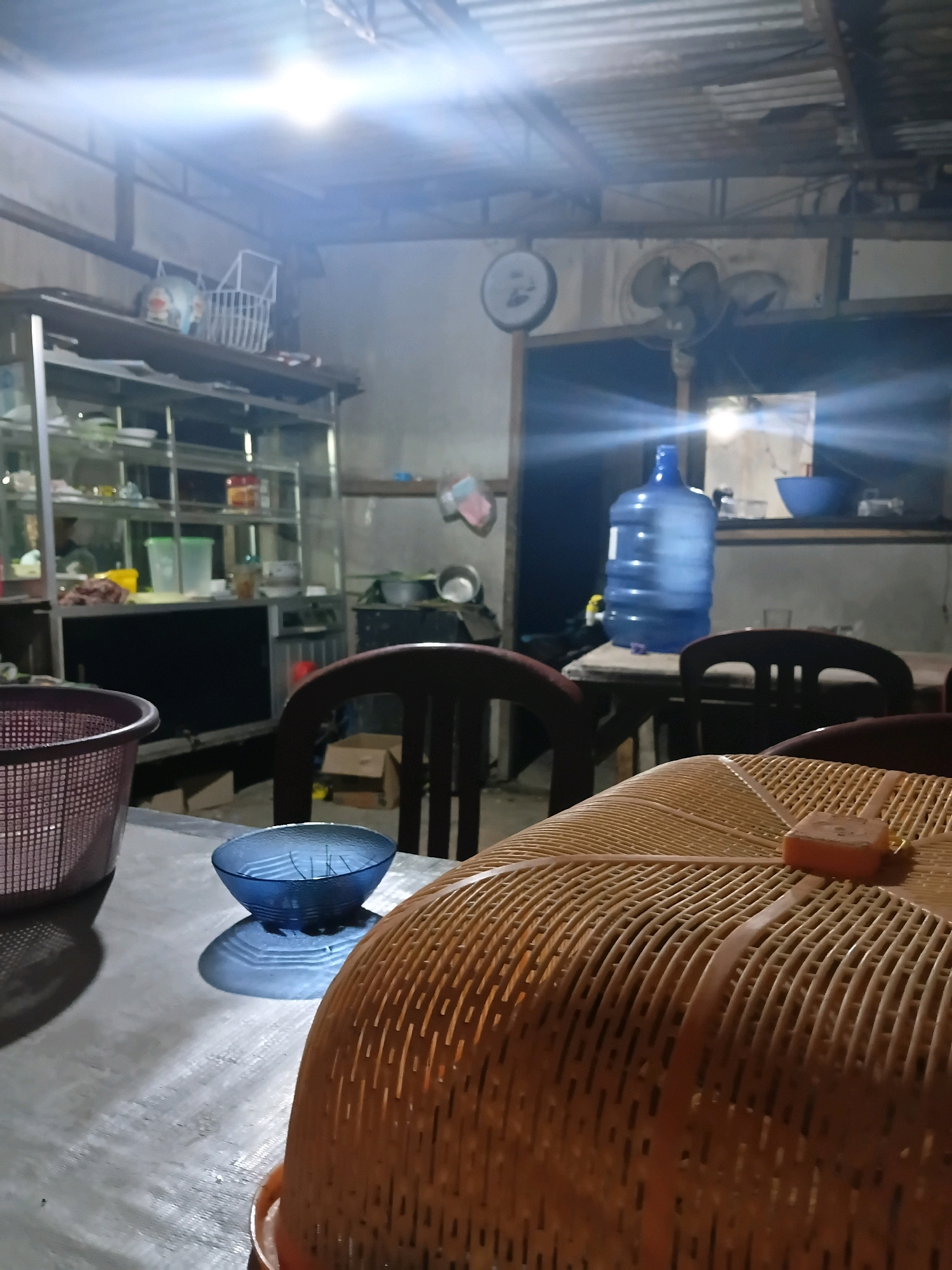 The image depicts the interior of what appears to be a casual eatery or home kitchen in Bantarbarang, Indonesia. The lighting is dim and primarily artificial, with a strong overhead light casting a bright glare. In the foreground, a metal table is visible, on which rests a blue glass bowl and part of a large, woven food cover. To the left, a pink woven basket is also present. Behind the table, several chairs are arranged, one of which is a dark wooden chair with a slatted back. Further back, shelves are filled with various items, including containers, and a large blue water jug stands prominently on a table. A wall fan is mounted on the wall, and a circular clock is visible above it. The overall environment suggests a functional, lived-in space. There are no people visible in the image.