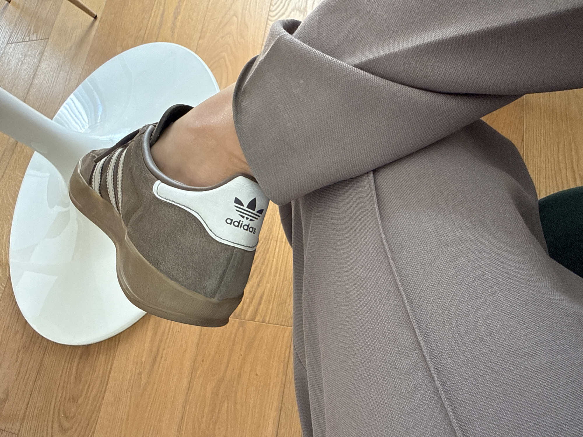 A close-up shot shows a person sitting with their right leg crossed over their left knee. The person is wearing gray trousers. On the right foot is a brown Adidas sneaker with white stripes and a white heel patch displaying the Adidas logo. The foot rests on a white plastic stool. The background consists of a wooden floor. The setting appears to be indoors. The location is not possible to determine.