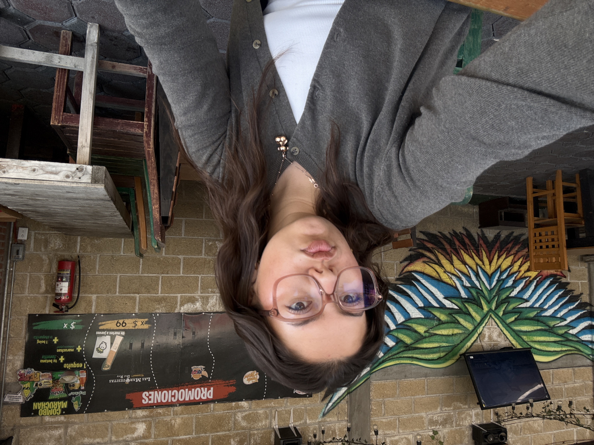 An image, rotated on its side, shows a young woman taking a selfie or portrait in what appears to be a casual eatery or market setting. The woman is centered in the frame, looking directly at the camera with a neutral expression and slightly pouted lips. She has dark, wavy hair, and wears large, translucent pink-framed glasses. She is dressed in a gray button-up cardigan over a white top, with a delicate gold necklace visible.
Behind her, a textured brick wall serves as the backdrop. To the viewer's left (her right), a red fire extinguisher is mounted on the wall. Below it, a large, dark promotional banner is displayed. Visible text on the banner includes "PROMOCIONES" (Promotions), "LAS MARQUESITAS Del Rey," and details about various food combos like "COMBO MARUCHAN" which includes "esquite," "TOSTITOS," "Pepelote," and offers like "$ x 66 de martes a jueves" (Tuesdays to Thursdays) and "$ x 1 boing o chaparrita + tu botana maruchan + tu botana."
To the viewer's right (her left), a vibrant, stylized mural depicting what looks like a bird or plant motif in blue, green, yellow, and red adorns the brick wall. Below the mural, a dark monitor or screen is mounted, and below that, a black speaker or audio device sits on a surface, accompanied by string lights. Various wooden chairs and tables are visible in the background, suggesting a dining area. The overall atmosphere is informal, possibly a local food stall or a relaxed restaurant in San Andrés Cholula, Mexico.