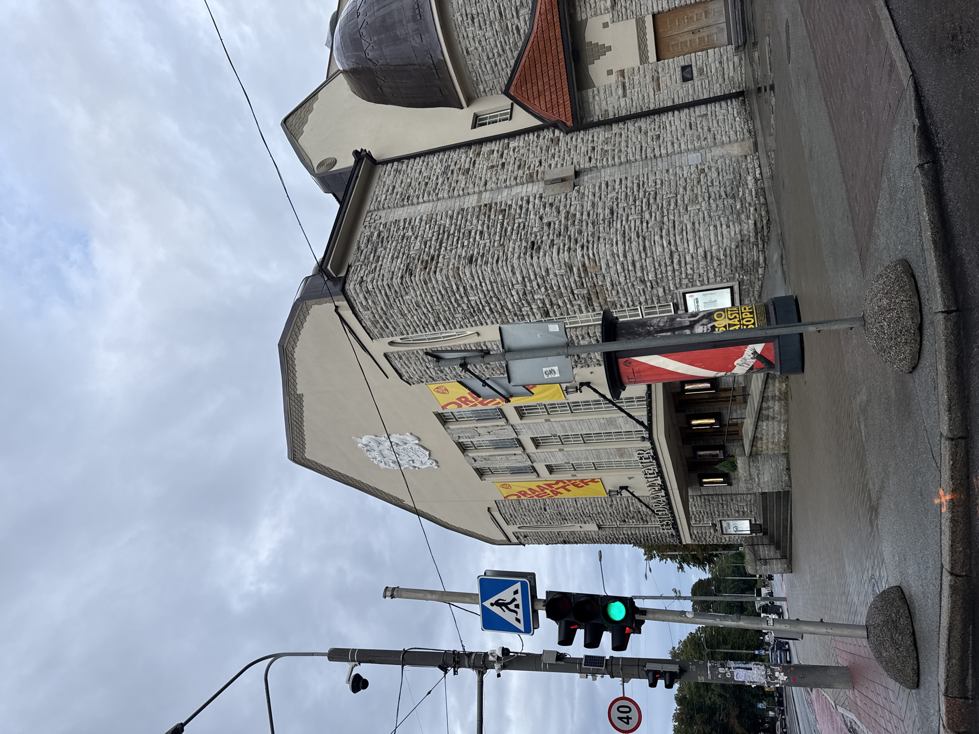 The image captures a view of the Estonian Drama Theatre in Tallinn, Estonia, on a cloudy day. The theatre's facade is predominantly made of stone, with large windows and a prominent white emblem above the entrance. Several yellow banners with text are displayed on the building. In the foreground, a traffic light shows a green signal, and a pedestrian crossing sign is visible. A speed limit sign indicating 40 is also present. The street and sidewalk appear wet, suggesting recent rain. The scene is captured from a street-level perspective, looking upwards towards the theatre and the sky.