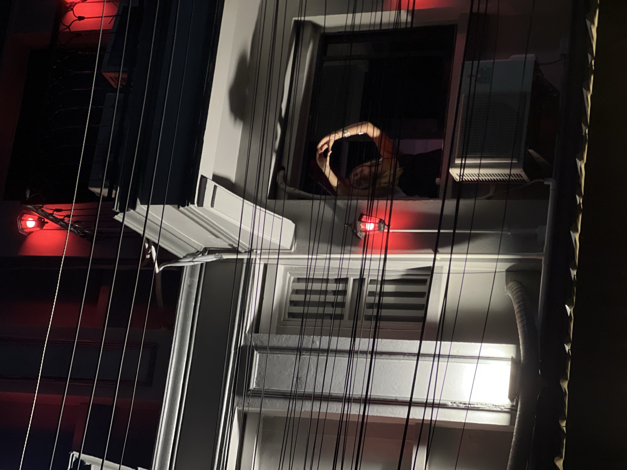 An outdoor shot features the side of a tall, light-colored building, possibly in a city. The view is from below, looking upward at an angle. The building's facade includes a window with a person seen inside, illuminated with interior light. The individual appears to be making a heart shape with their hands. To the right of the window is an air conditioning unit protruding from the building. Additionally, there is a window with blinds that are closed. The building is decorated with strands of lights, some of which are red. There is a vent in the lower right corner. The city location is unknown.