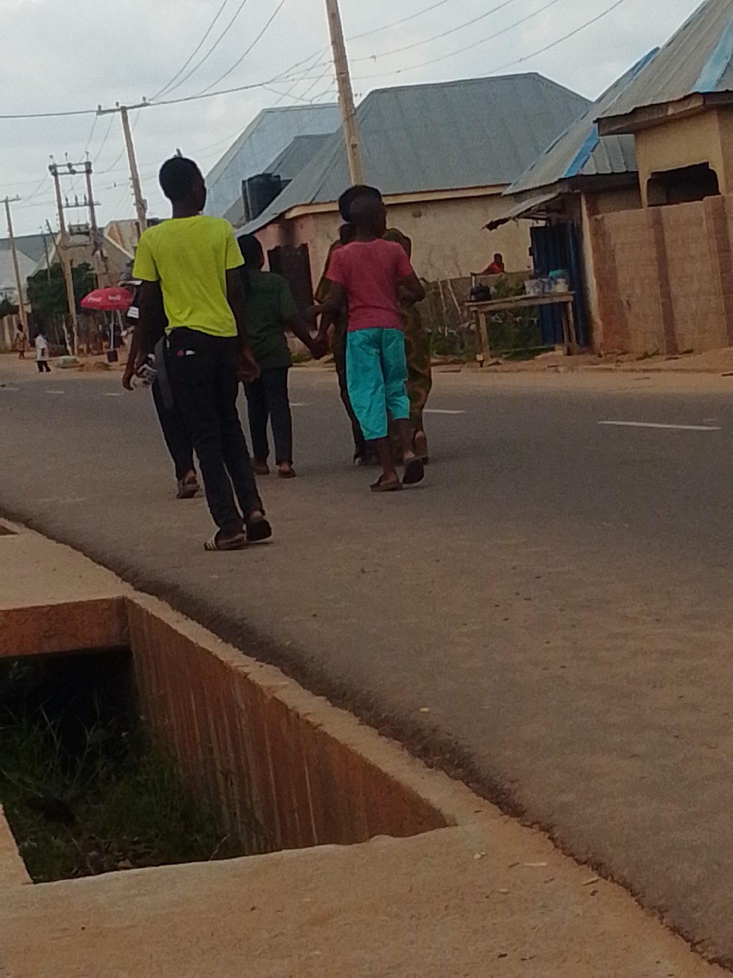 The image captures a street scene in Gombe, Nigeria, under an overcast sky. In the foreground, a concrete drainage ditch runs along the left side of the road. The road itself is asphalt with a white line marking the center. Several people are walking on the road. On the left, a person wearing a bright green t-shirt and black pants walks away from the camera. To their right, another person in a dark green shirt and dark pants is also walking away. In the center, a person in a pink t-shirt and turquoise shorts walks forward, with their back to the camera, holding hands with someone behind them. Further back, other individuals and structures are visible, including buildings with corrugated metal roofs, a red umbrella, and utility poles with power lines. The general atmosphere appears to be that of a typical day in a Nigerian city.