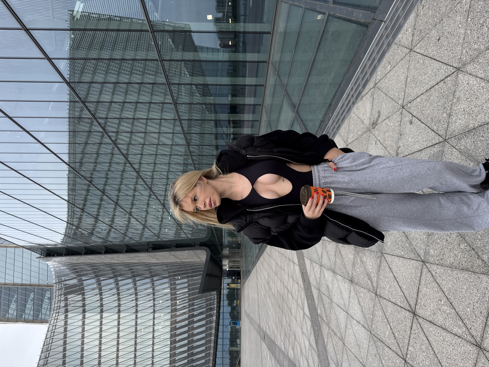 A blonde woman in a black cutout bodysuit, a black puffer jacket, and grey sweatpants holds an orange coffee cup in her right hand. She is standing on a paved plaza in front of a modern glass building. The building's facade is composed of numerous geometric panes of glass, creating a reflective surface that mirrors the sky and surrounding urban environment. The plaza pavement consists of large, grey rectangular tiles arranged in a repeating geometric pattern. The overall impression is one of contemporary urban architecture in an outdoor setting. The location is identified as Saint Petersburg, Russia, suggesting this is likely a business district or a public space within the city.