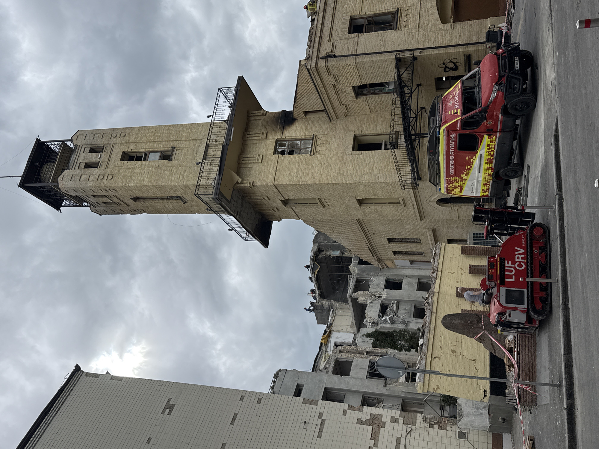 The image shows a building in Kyiv, Ukraine, severely damaged by an apparent bombing or shelling. A tall tower, largely intact but with broken windows, stands beside a building reduced to rubble. Rescue workers are visible on top of the damaged structure. A red emergency vehicle and a smaller tracked vehicle are parked on the street in front of the buildings. There is safety tape marking off the area. The sky is overcast.