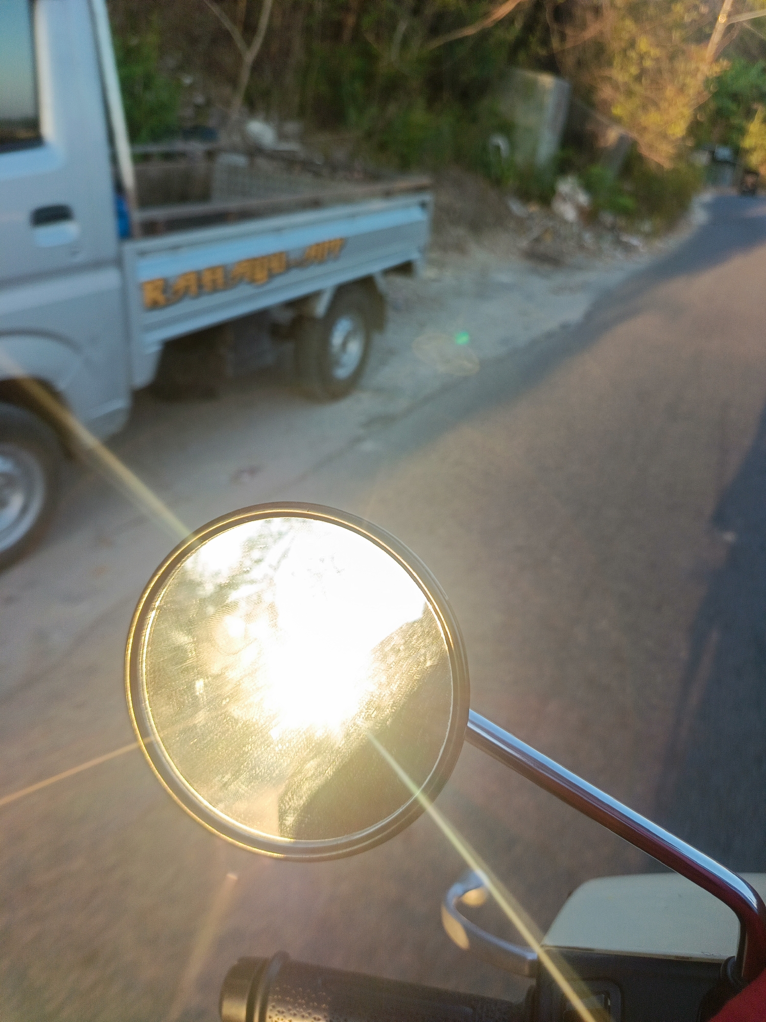 The image shows a side-view mirror of a vehicle, reflecting the bright sun and a dusty road. In the background, a light-colored pickup truck with orange lettering on its side is visible, parked on a dirt shoulder next to the road. The environment appears to be rural or semi-urban, with vegetation and some debris on the roadside. The lighting suggests it is either sunrise or sunset, casting long shadows. The scene is likely in Gorontalo, Indonesia, given the context provided.