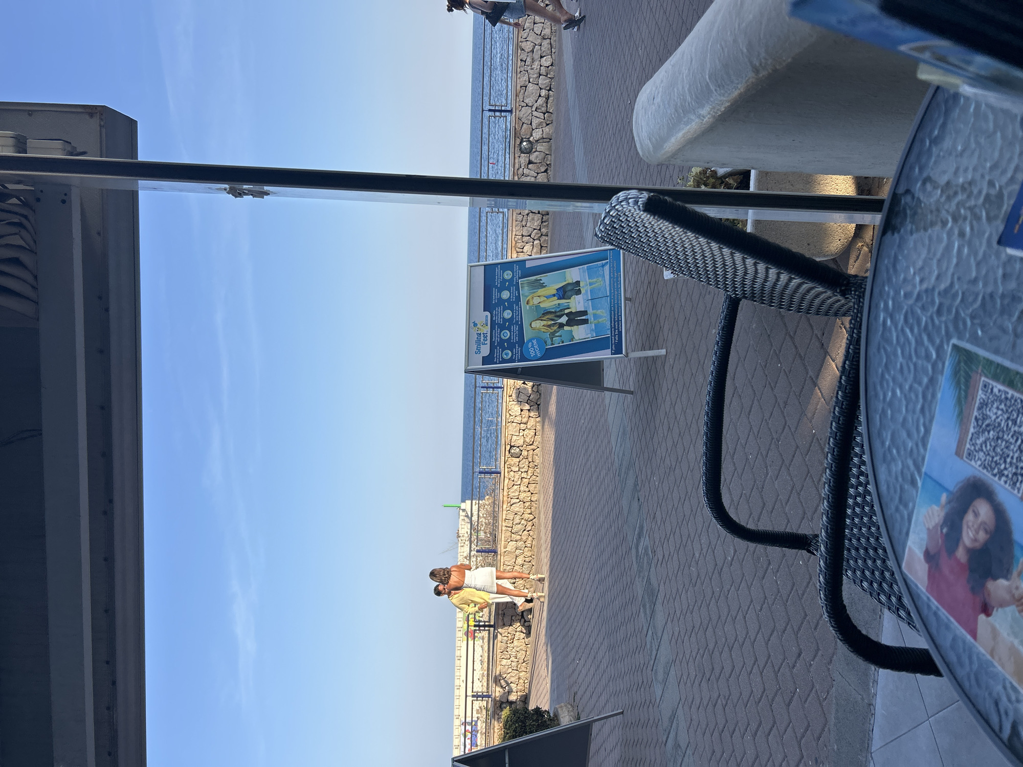 The image depicts a view from a covered outdoor seating area, likely a cafe or restaurant, overlooking a paved walkway and the sea. In the mid-ground, two individuals are walking along the walkway near a stone wall that separates it from the sea. A sign with graphics and text, possibly advertising a service, stands on the walkway. Beyond the walkway, the calm blue sea stretches to the horizon under a clear sky. In the foreground, a glass-topped table with a wicker chair is visible, with some papers or brochures on the table. The overall ambiance suggests a pleasant, sunny day in a coastal town. This scene is located in Cala Rajada, Spain, a coastal resort town on the island of Mallorca.