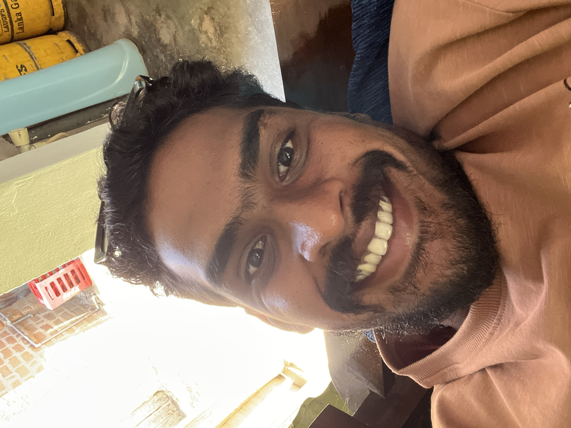 A close-up selfie of a smiling man with dark hair, a beard, and brown skin. He wears a light pink or tan long-sleeved shirt. In the background, there is a yellow gas canister with the word "LAUGFS" visible. Behind the man, there is a blue object. A Coca-Cola bottle carrier sits on a brick surface in the left background. The location is Unknown, Unknown.
