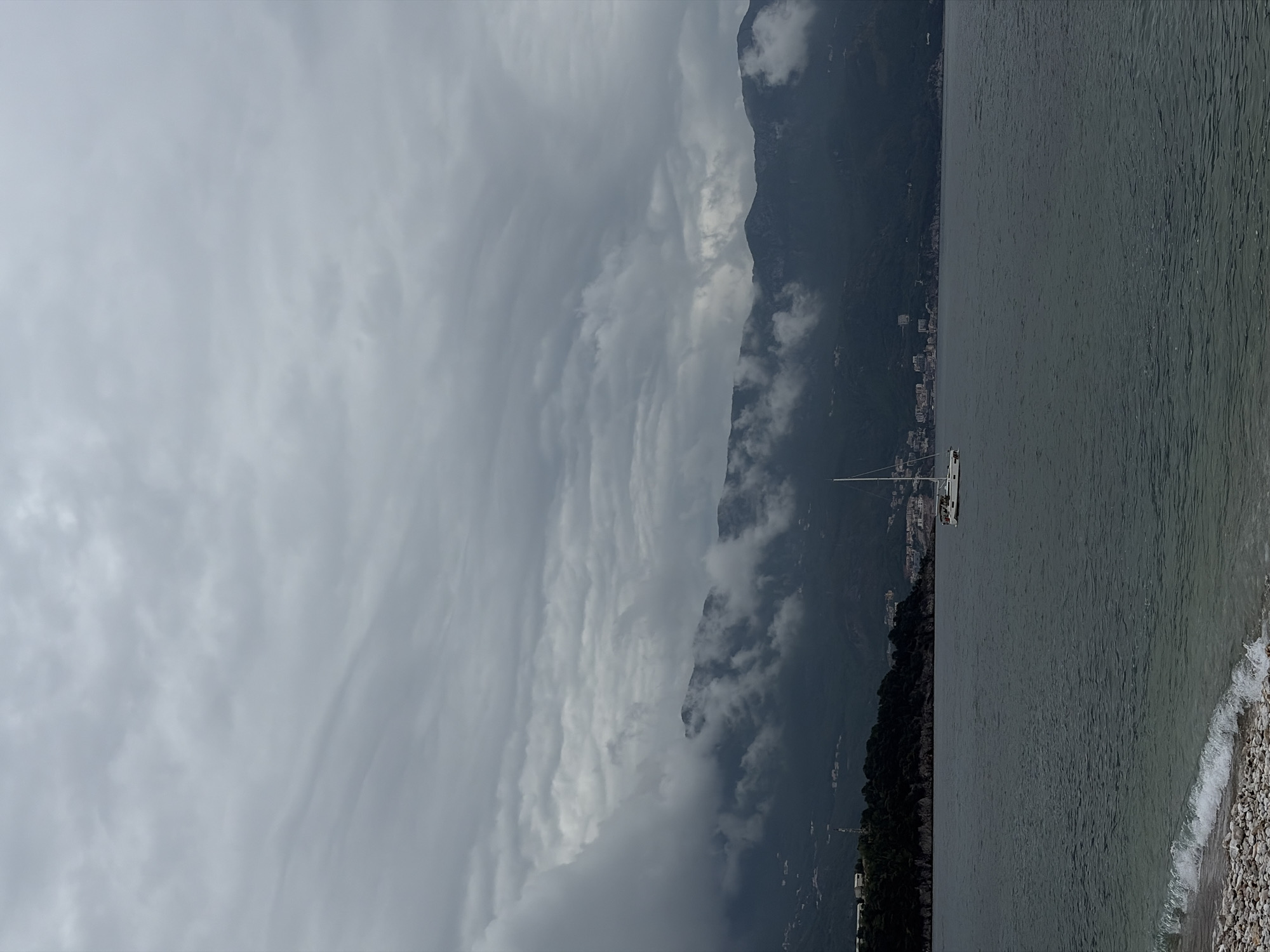 The image captures a scenic view in Budva, Montenegro. Dominating the scene is a mountain range, partially obscured by clouds. Along the coast, there is a coastline with a pebble beach. A sailboat is anchored in the water off the coast. The sky is overcast. Buildings are visible along the mountainside.