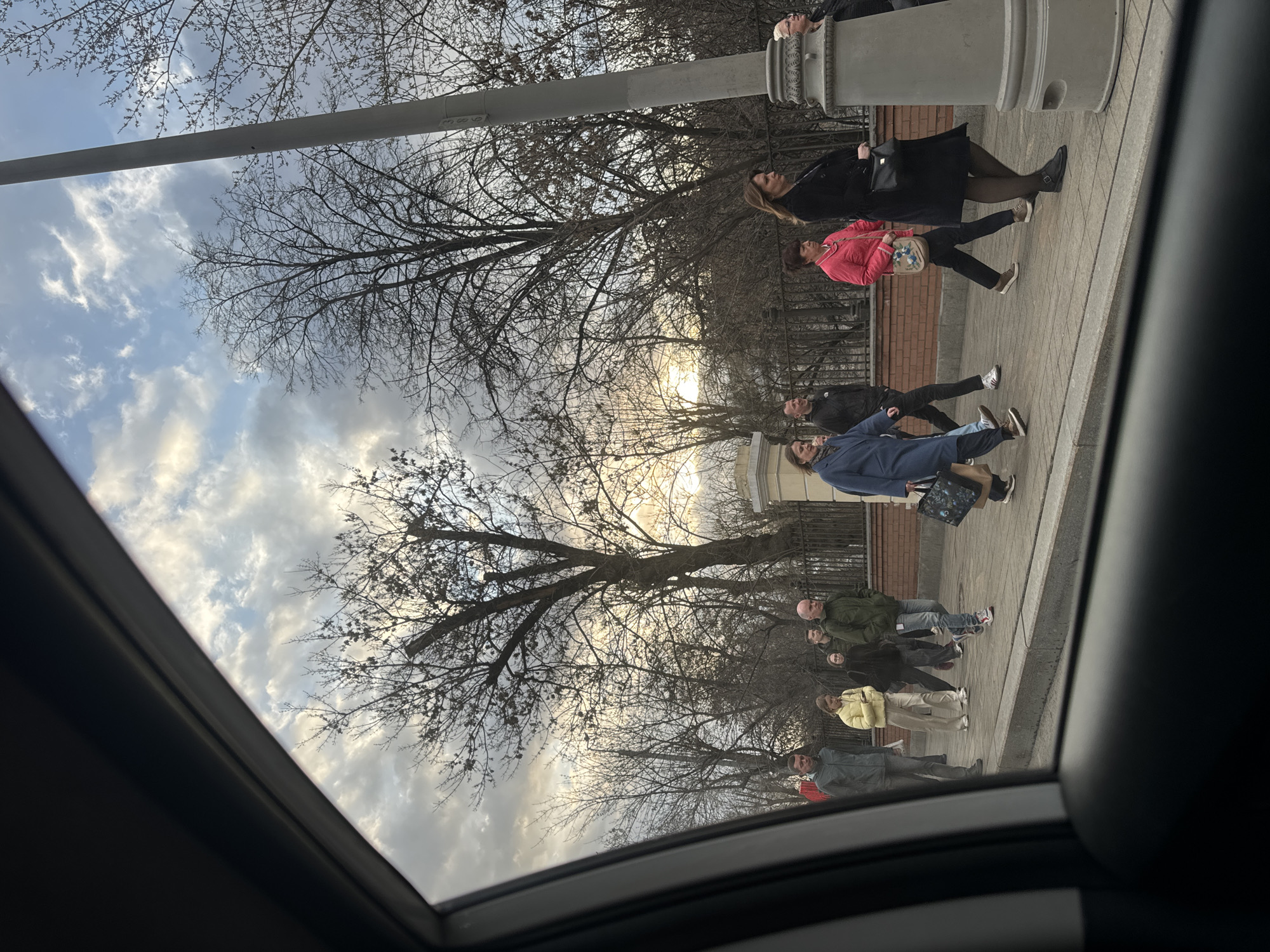 A photograph taken from inside a vehicle shows a group of pedestrians walking along a sidewalk near a brick building and a decorative wrought-iron fence. Leafless trees are visible, and the sky is partly cloudy. The location is Moscow, Russia.