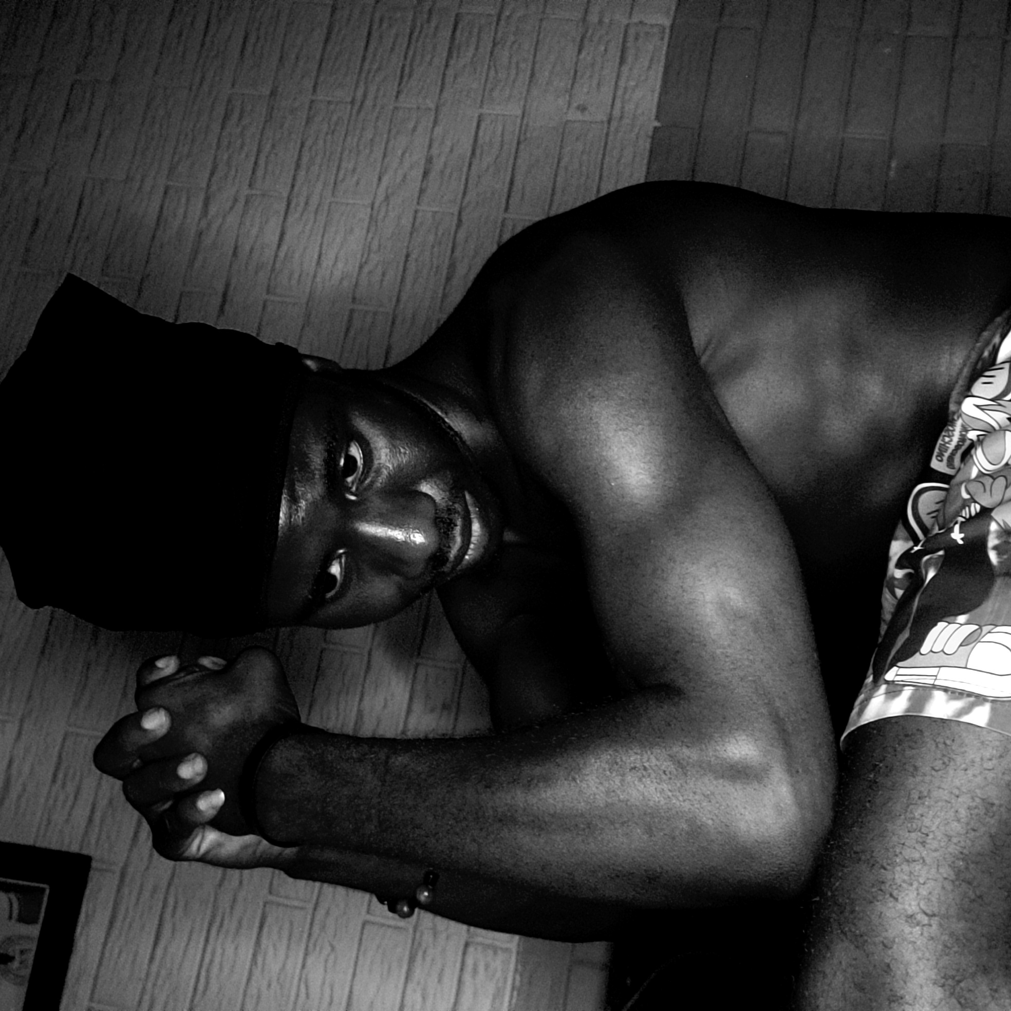 A shirtless black man wearing a black head covering is kneeling on the floor. He is facing forward with his hands clasped together near the corner of a wall, with a small portion of a picture frame visible. The man is wearing patterned shorts. The background appears to be a wall with rectangular tiles. The image is in black and white, and the location is in Narayi, Nigeria.