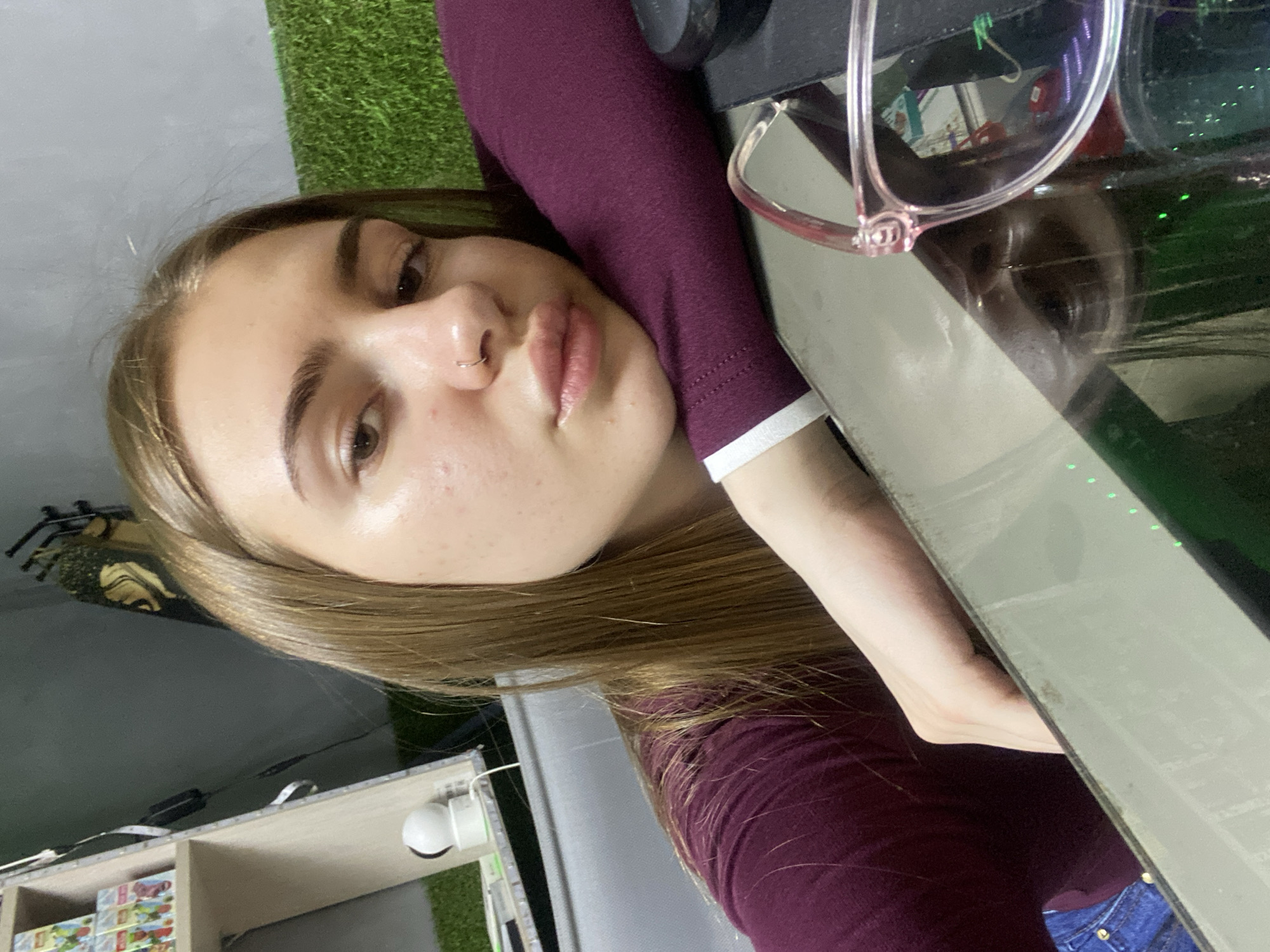 A young woman with long, light brown hair and a nose ring is leaning on a glass surface, with her right arm resting on a purple long-sleeved shirt. She is looking towards the camera. In the background, there is a shelf with various items, a white security camera, and a green textured wall. A pair of pink-rimmed glasses is visible in the foreground. The location is specified as Dyakovo, Russia, though the visual elements do not provide specific clues to confirm this location.