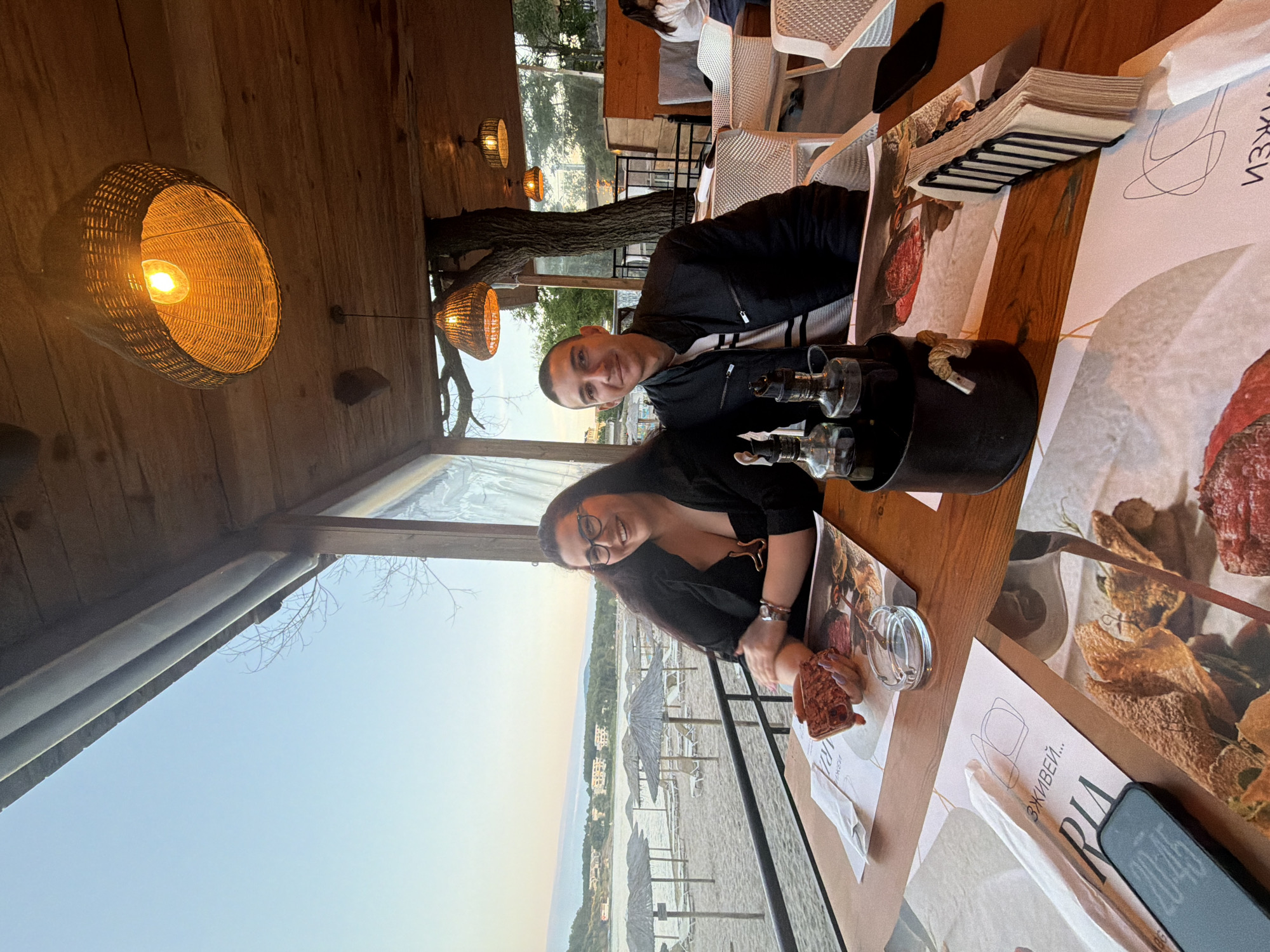 A man and a woman are seated at a wooden table on a covered outdoor patio overlooking a beach in Tsarevo, Bulgaria. The woman is on the left, wearing glasses and a dark top, and is holding up a small item that appears to be a piece of food. The man is on the right, wearing a dark jacket over a striped shirt. In front of them are menus, a napkin holder, an ashtray, and a set of glass condiment bottles. In the background, beach umbrellas and loungers are visible on the sand, and buildings are scattered along the coastline under a pale sky. The patio has a wooden roof with decorative pendant lights.