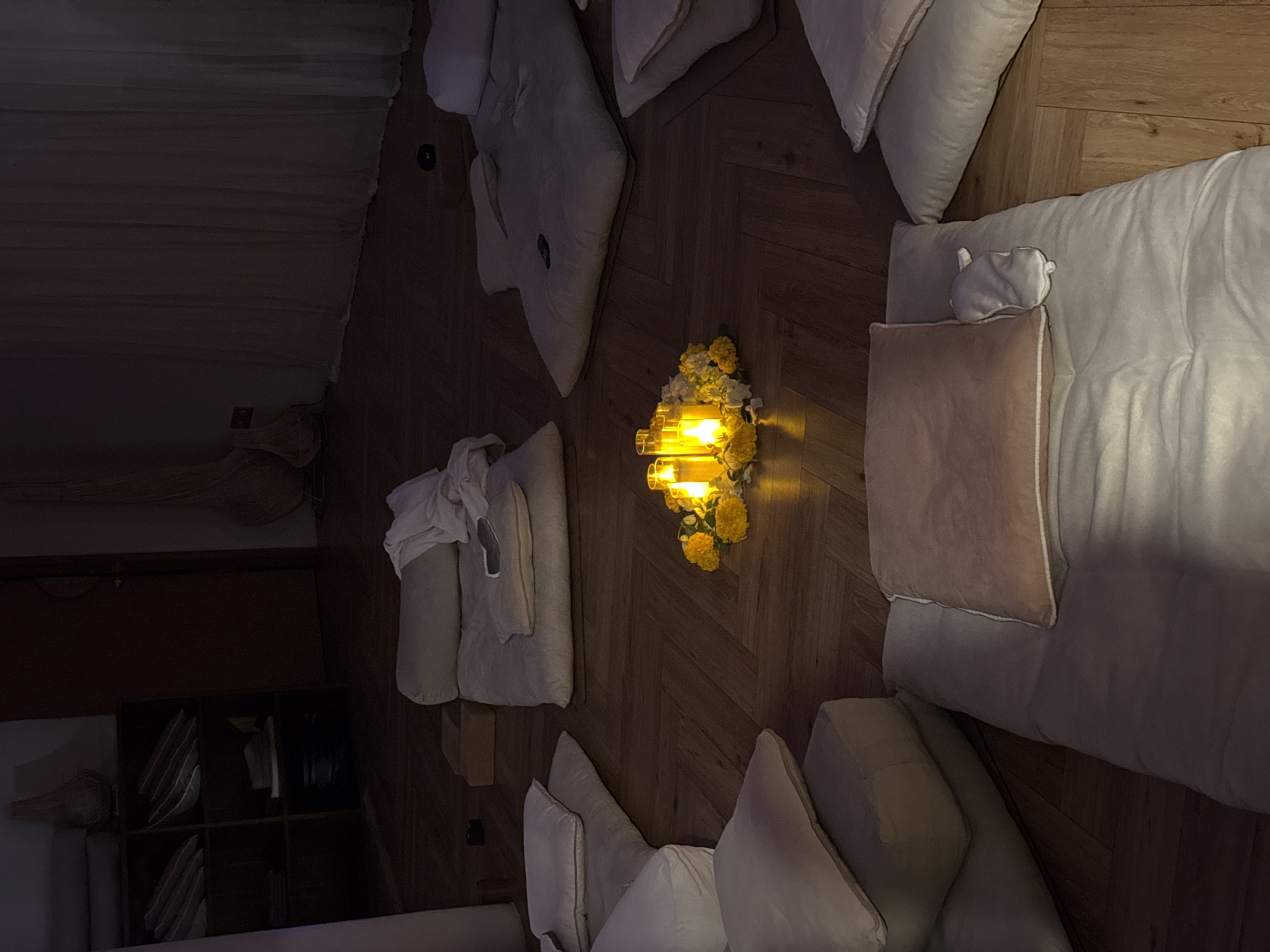 In a dimly lit room with wooden flooring, several floor cushions and futons are arranged in a circular fashion. At the center of this arrangement, a cluster of yellow flowers encircles three lit pillar candles, casting a warm glow. The room appears to be set up for a meditation or relaxation session. In the background, a dark wooden shelving unit is visible, stocked with folded items, and a closed door is present. The overall ambiance is serene and tranquil, suggesting a private indoor space, possibly in a retreat or wellness center in Mengwi, Indonesia. There are no people or animals visible in the image.